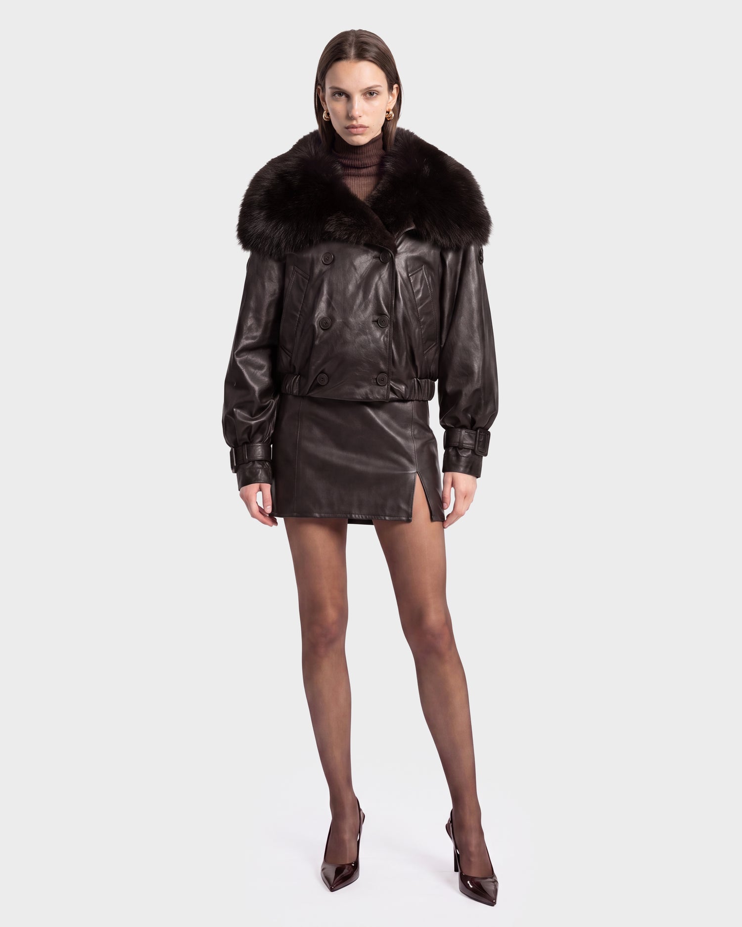 Styled in the Rudsak JANELLE - 8126936 Espresso, this woman showcases a bold modern look with a dark leather bomber jacket featuring a statement faux fur collar, paired with a matching leather mini skirt with a front slit, sheer tights, and pointed heels—all set against a plain light background for maximum impact.