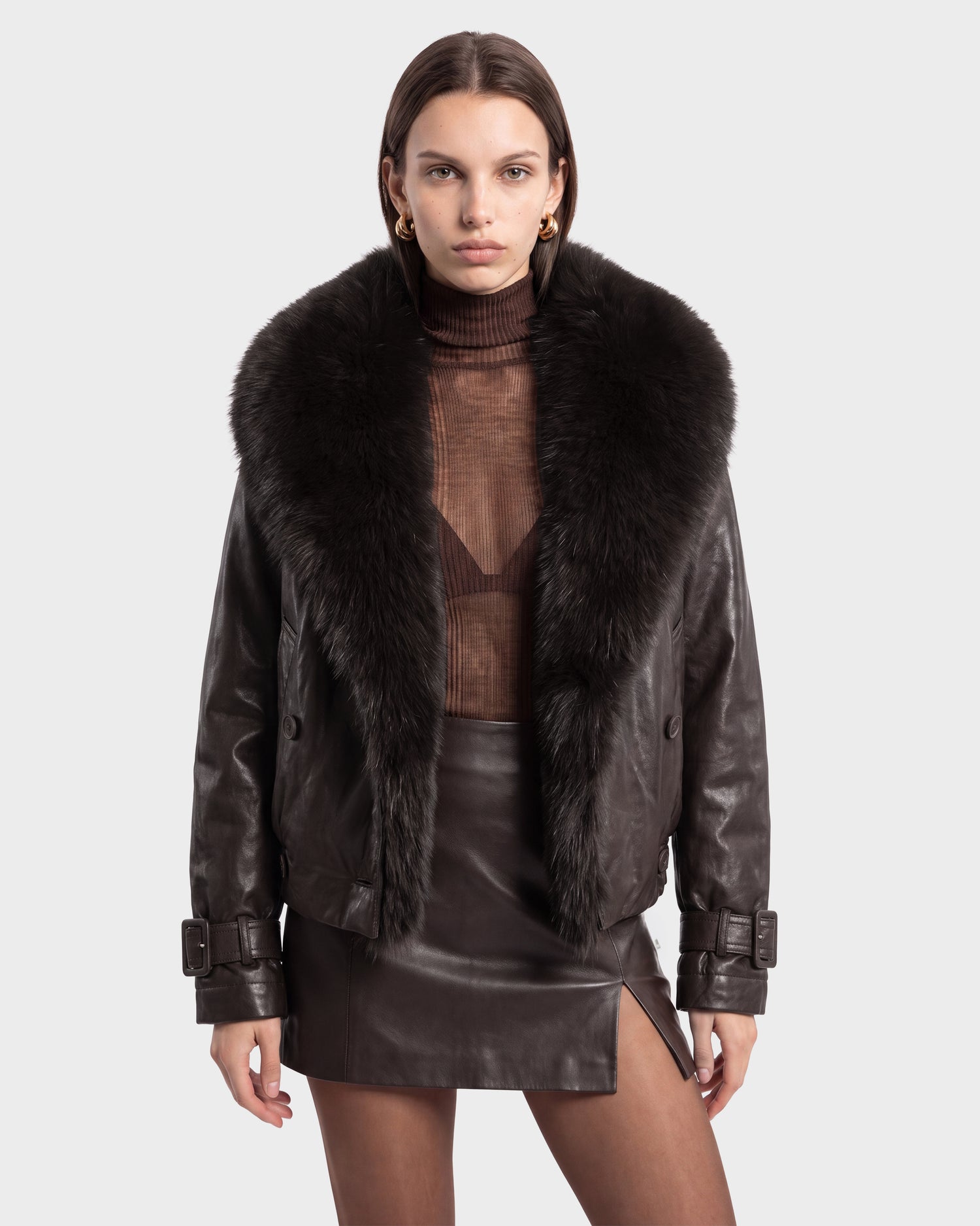A woman stands against a plain white background wearing the Rudsak JANELLE - 8126936 Espresso bomber jacket, featuring black leather and a statement faux fur collar. She pairs it with a sheer brown turtleneck top and a black leather mini skirt with a front slit, creating an ensemble that effortlessly blends edge and sophistication with a modern silhouette.