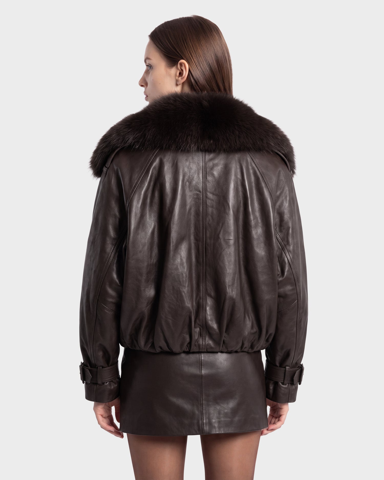A woman seen from behind wears the Rudsak JANELLE - 8126936 Espresso dark brown leather bomber jacket featuring a large faux fur collar, paired with a matching leather mini skirt, set against a plain light background to showcase its modern silhouette.