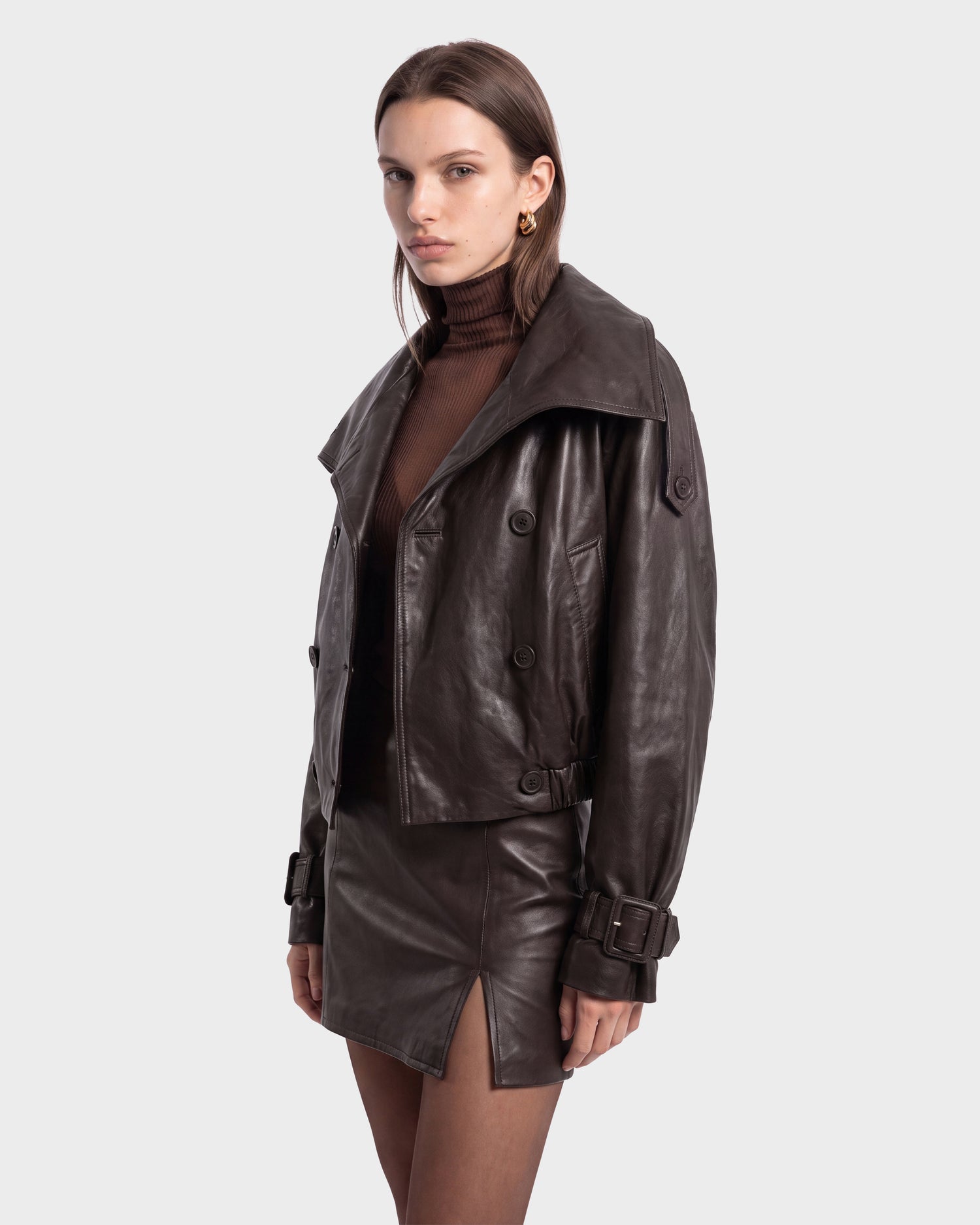 A woman wearing the Rudsak JANELLE - 8126936 Espresso dark brown leather bomber jacket with a modern silhouette and matching skirt featuring a front slit, paired with a brown turtleneck top, stands against a plain white background.