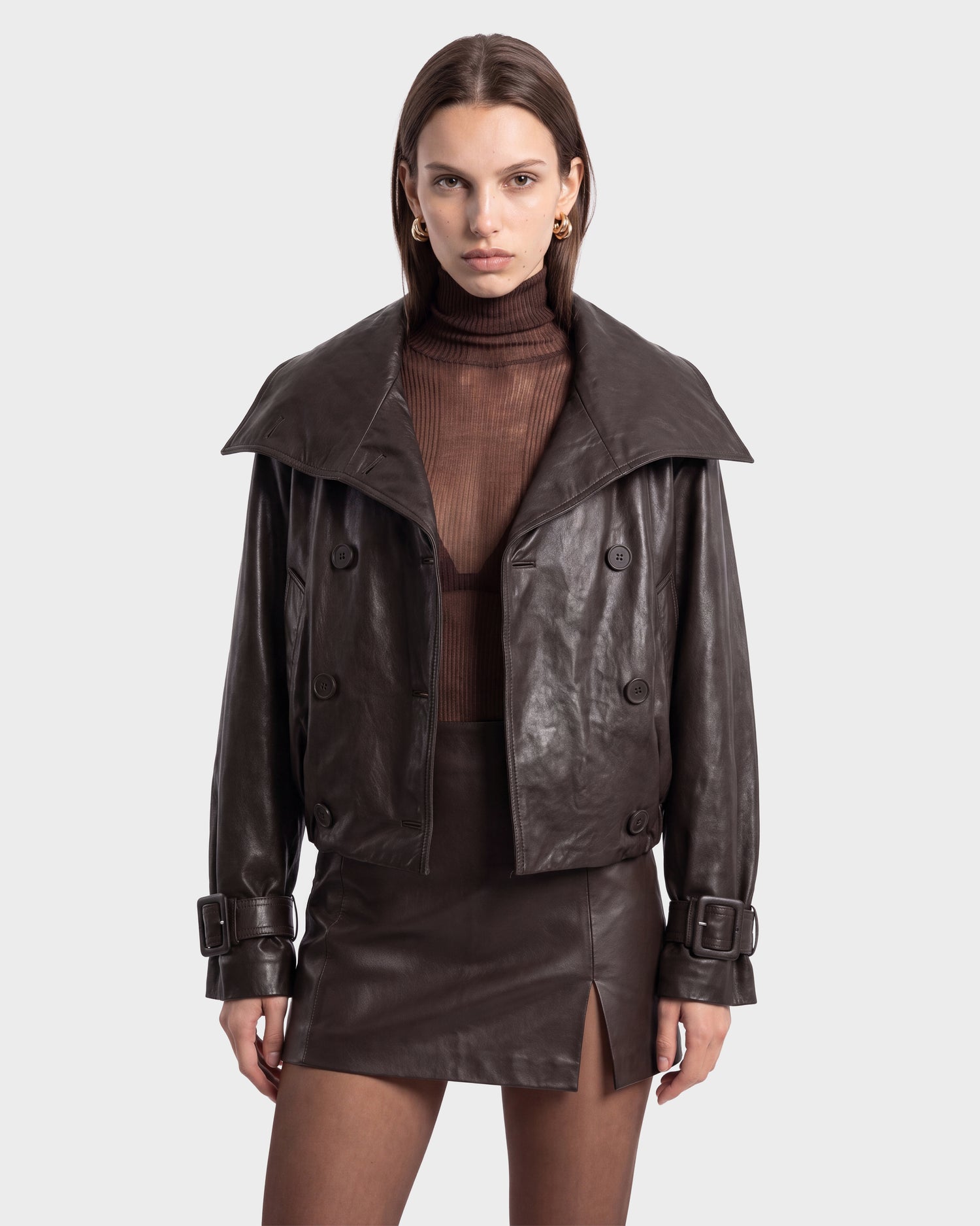 A woman stands facing forward against a plain background, wearing the JANELLE - 8126936 Espresso brown leather bomber jacket by Rudsak, styled with a modern silhouette, over a brown sheer turtleneck and a brown leather mini skirt.