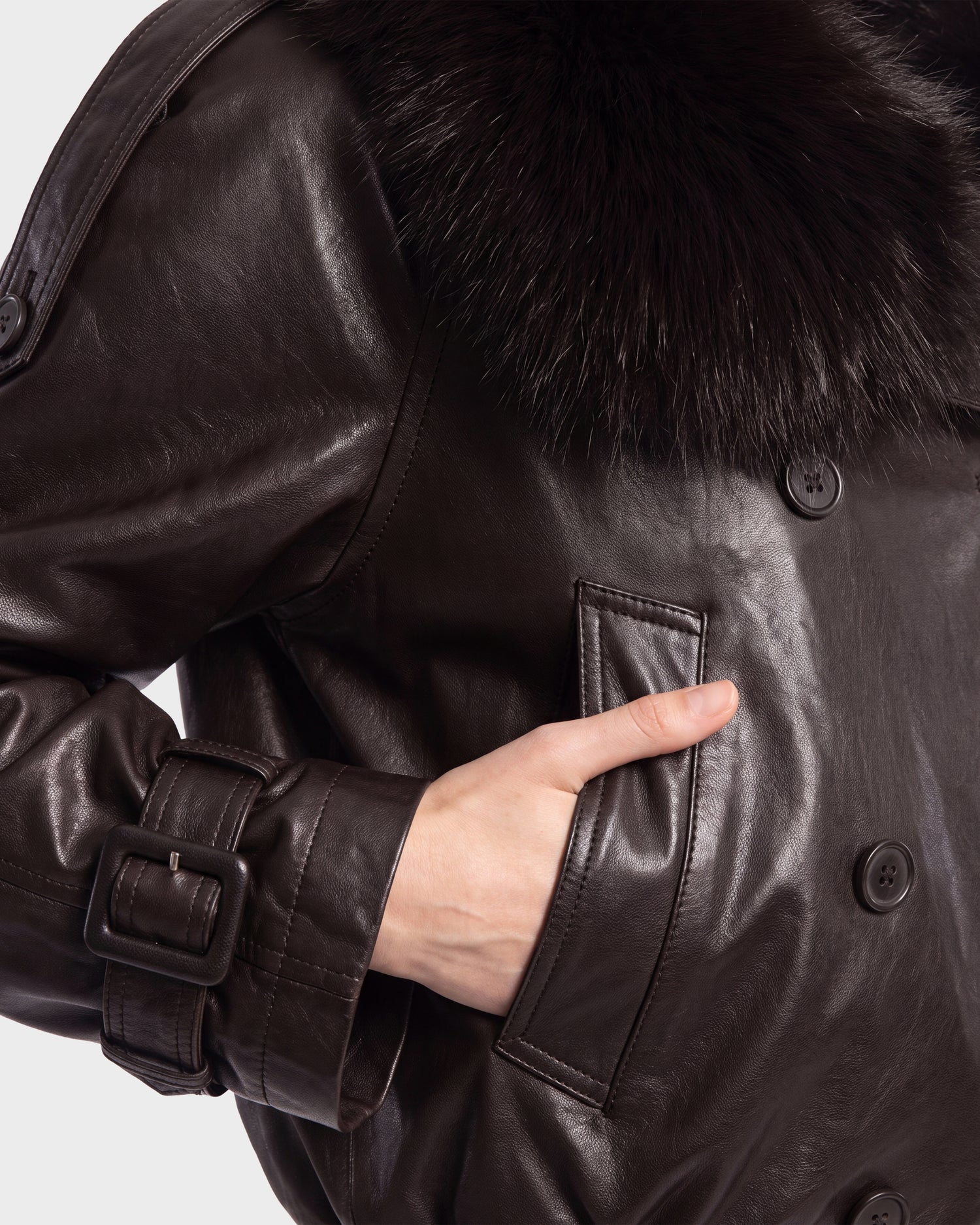 A person is wearing the Rudsak JANELLE - 8126936 Espresso, a dark brown leather bomber coat with a faux fur collar. The design features large buttons, a belt with a buckle on the sleeve, and side pockets, creating a modern silhouette.