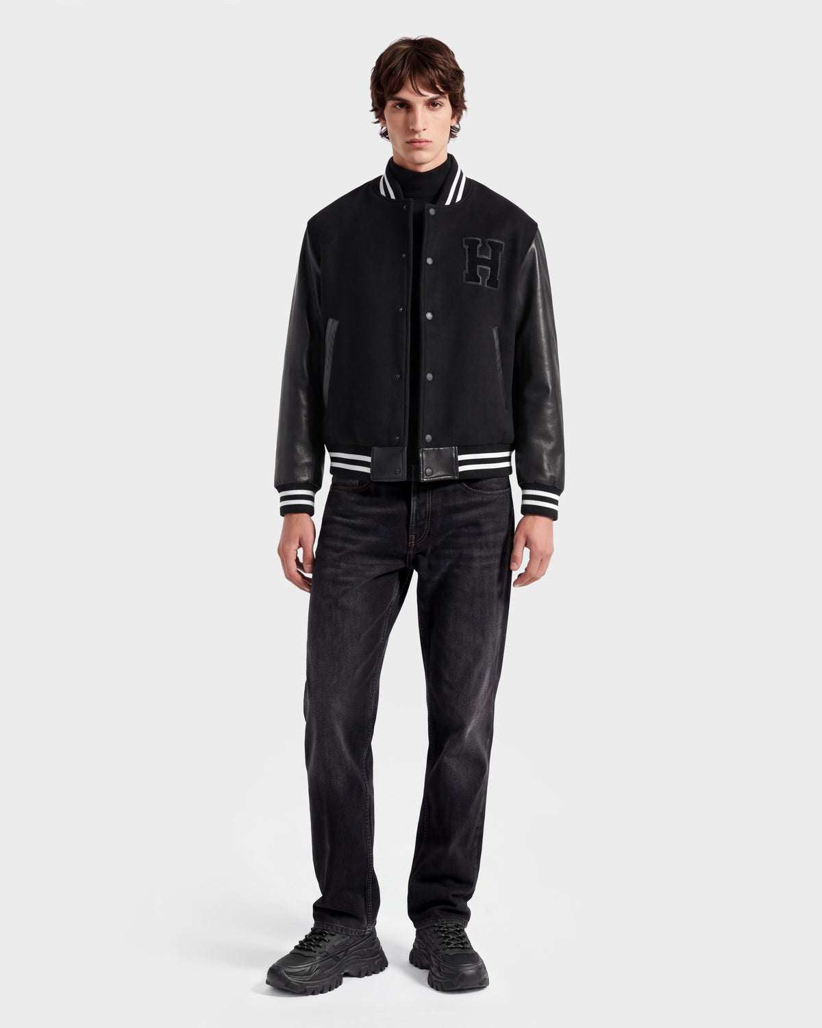 A young man in a limited-edition Harvard varsity jacket with leather sleeves, Rudsak JOHN HV - H125504 Black jeans, and black sneakers stands facing forward against a plain white background, channeling classic Ivy League style.