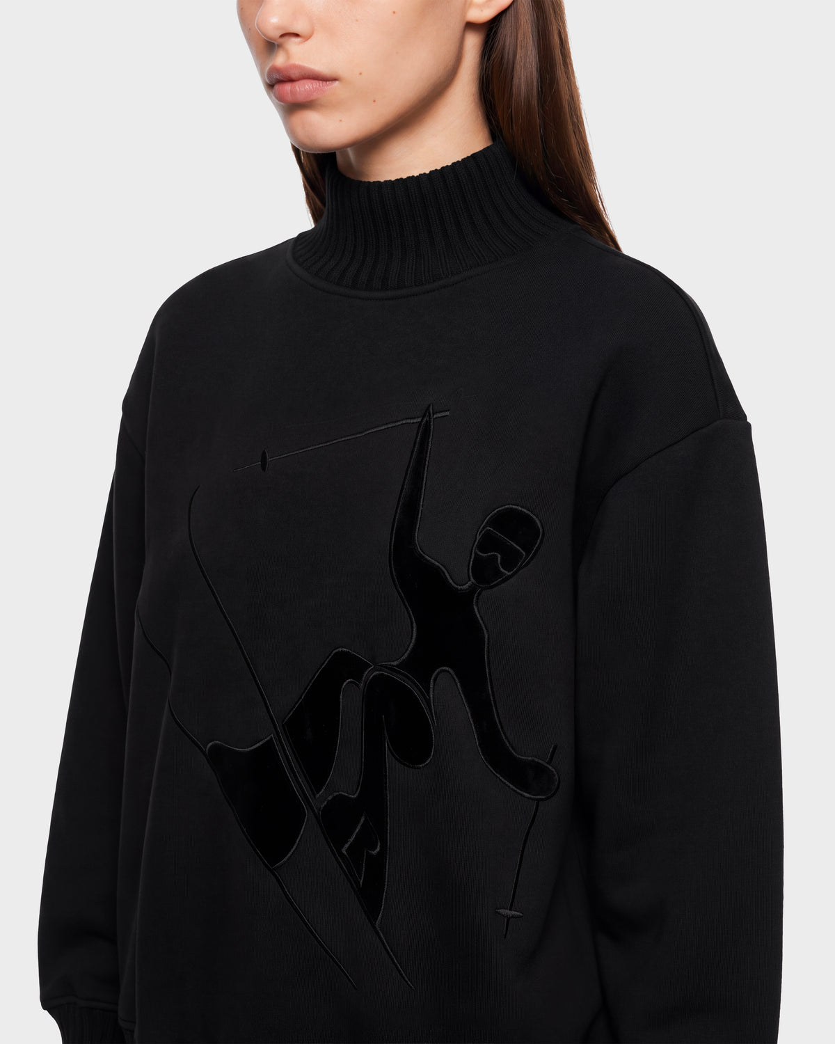A woman wearing the Rudsak JOYCE E - 8526538 Black turtleneck sweater, featuring an embossed abstract skier design on the front, photographed from the shoulders up against a plain light background.