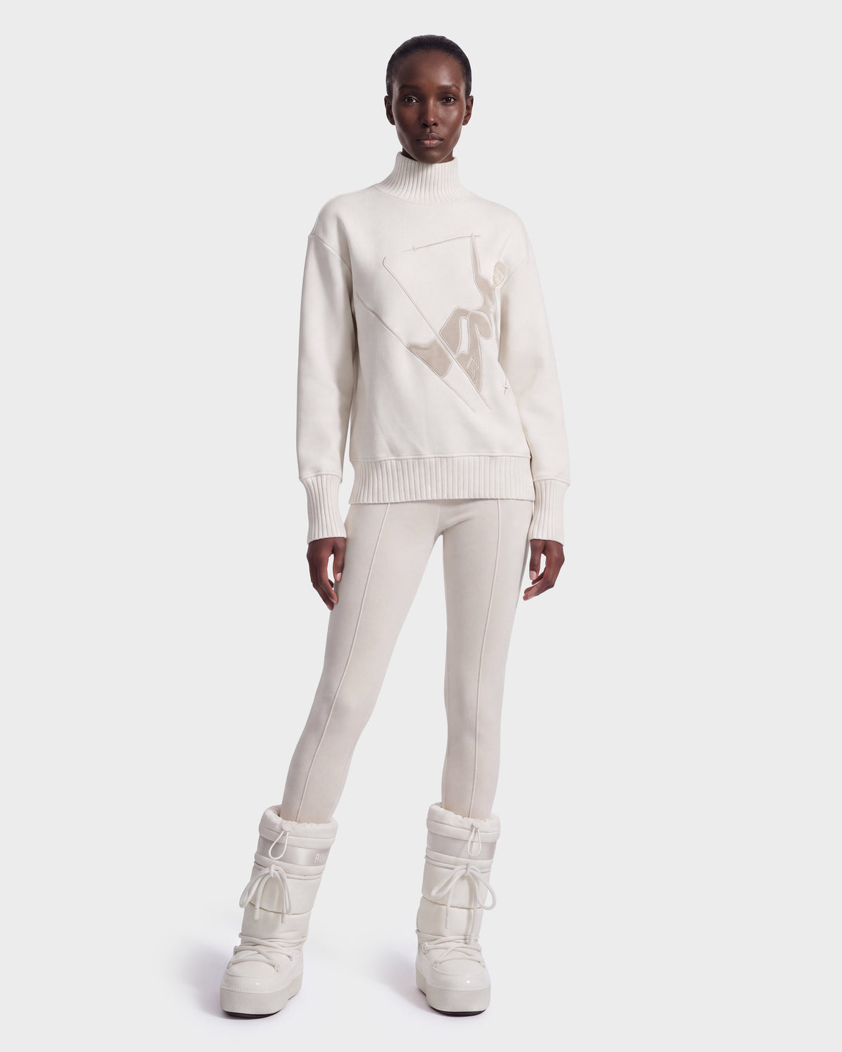 A person stands facing forward against a plain white background, wearing the Rudsak JOYCE E - 8526538 Vanilla chunky cream snow boots, paired with a cream turtleneck sweater and matching fitted pants.