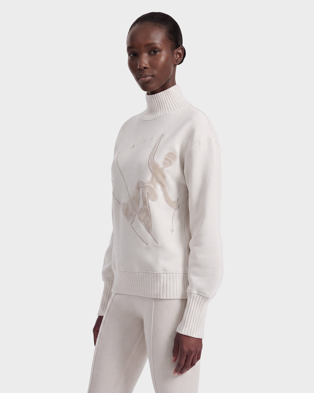A woman wearing the Rudsak JOYCE E - 8526538 Vanilla turtleneck sweater with an abstract figure design and matching white pants stands against a plain background, looking slightly to the side.