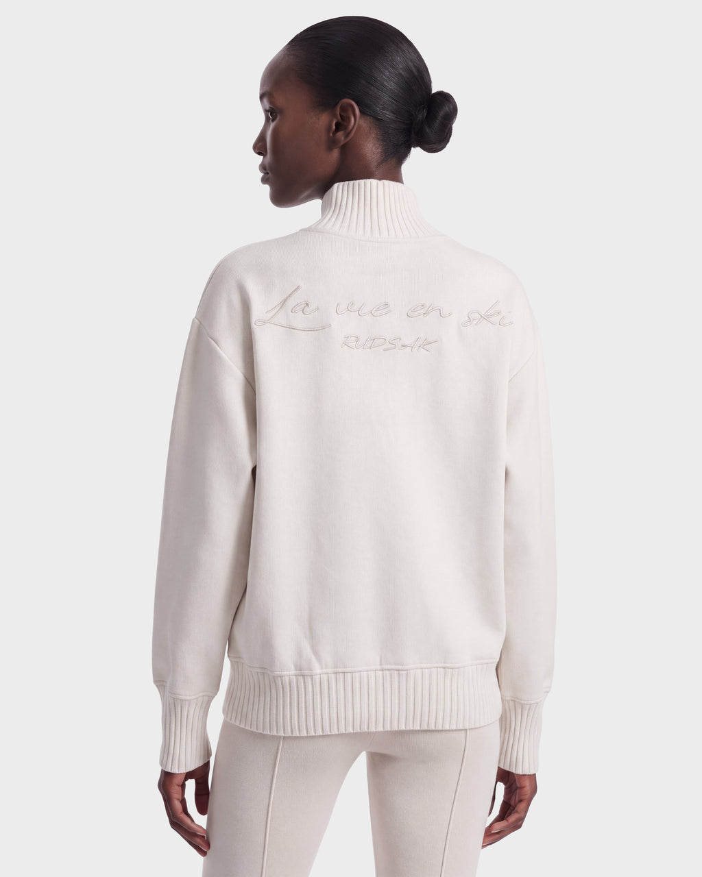 A woman stands facing away, hands by her sides, wearing the Rudsak JOYCE E - 8526538 Vanilla off-white turtleneck sweater with “La vie en ski” embroidered on the back.