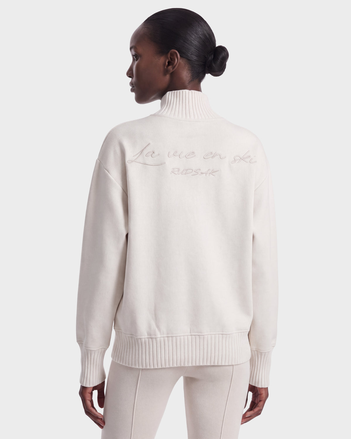 A woman stands facing away, hands by her sides, wearing the Rudsak JOYCE E - 8526538 Vanilla off-white turtleneck sweater with “La vie en ski” embroidered on the back.