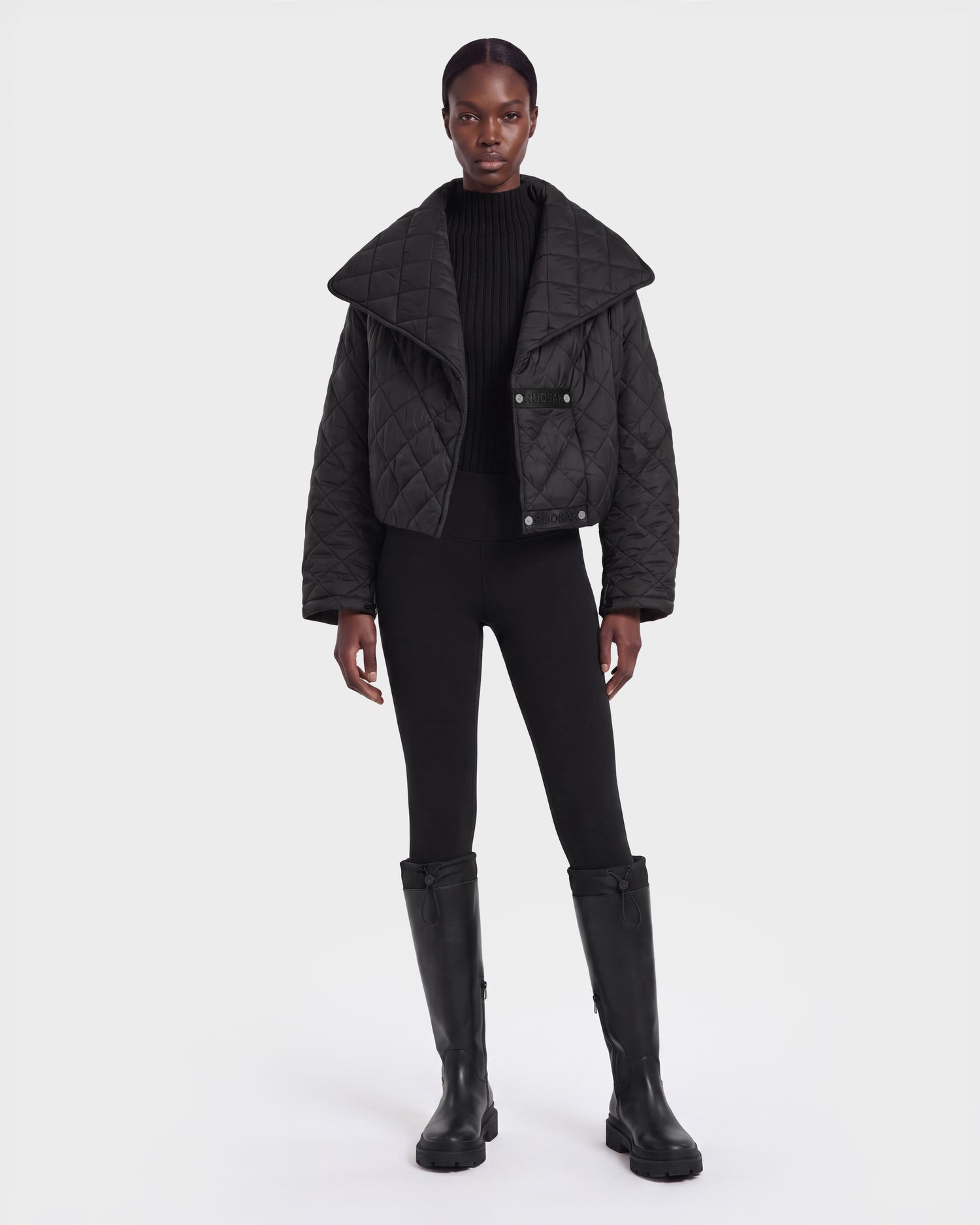 A person stands facing forward, wearing the Rudsak KALEE - 8124005 Black women’s quilted light jacket with a wide collar, paired with a black sweater, black fitted pants, and tall black boots, against a plain white background.