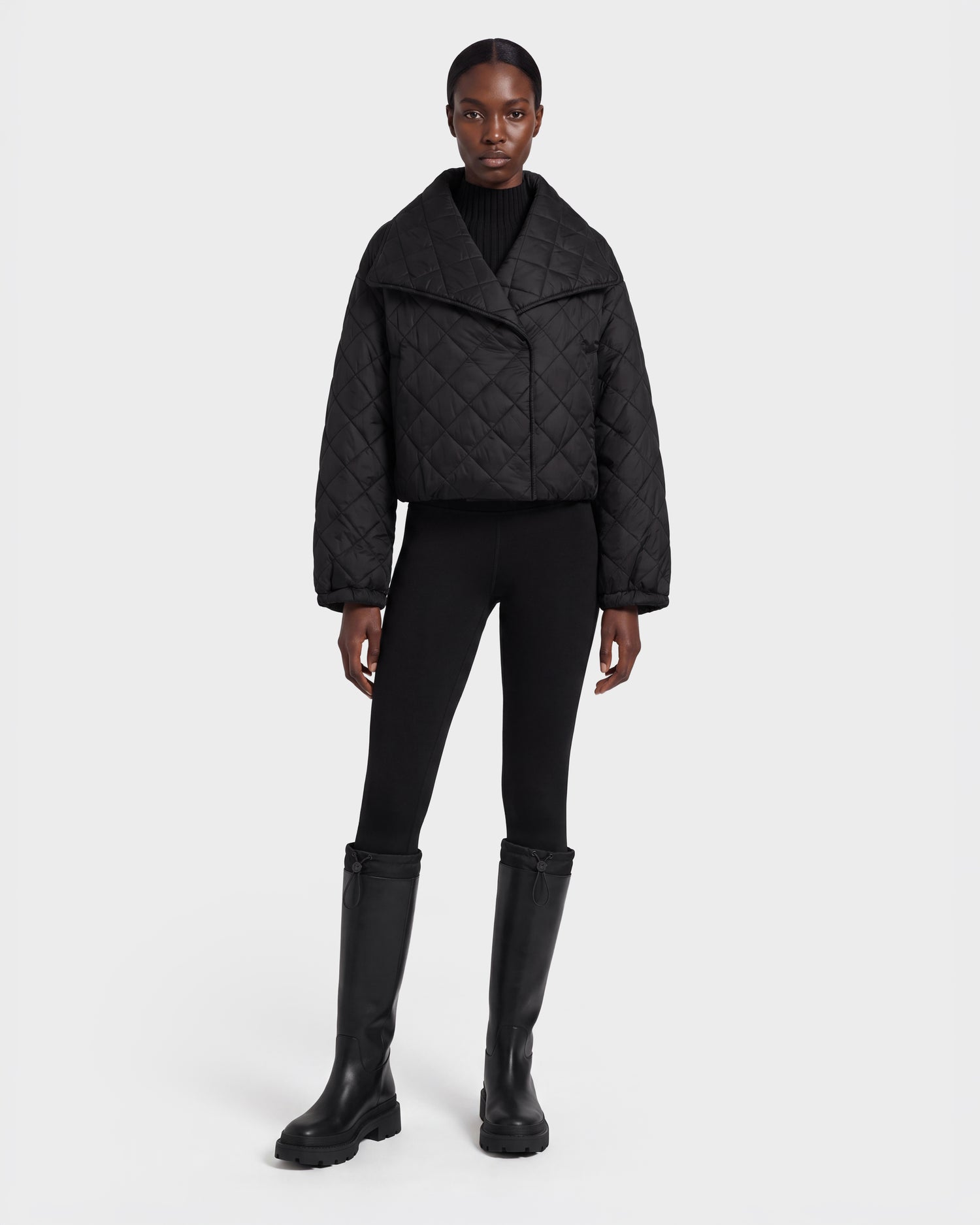 A person stands facing forward, wearing the Rudsak KALEE - 8124005 Black women's quilted light jacket styled for active versatility, paired with black leggings and knee-high black boots against a plain white background.