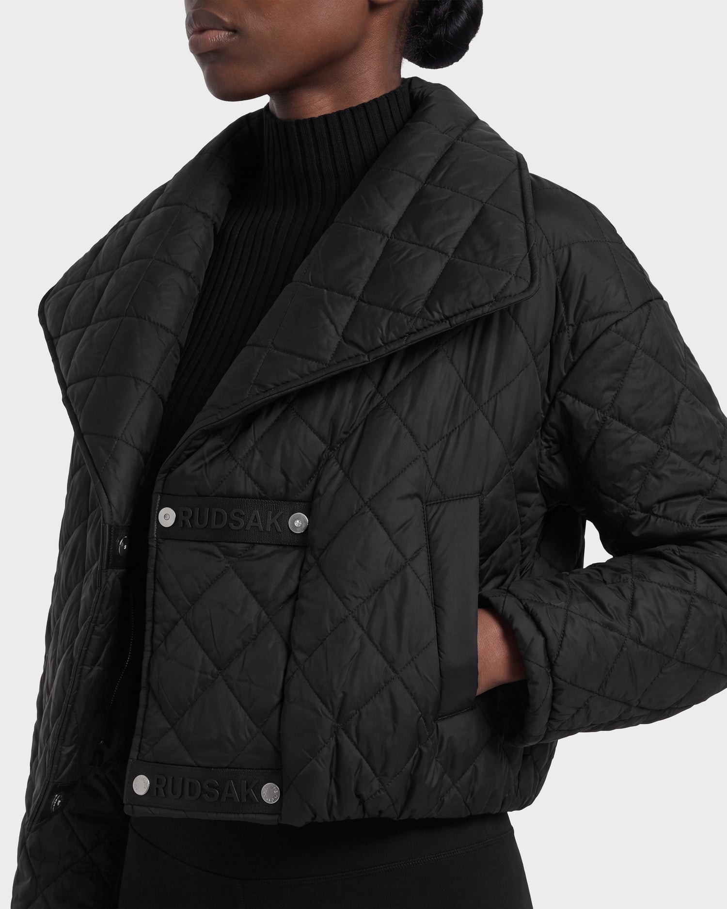 A person wearing the KALEE - 8124005 Black women's quilted light jacket by Rudsak, featuring wide lapels, snap buttons, and side pockets with their hands tucked in. The jacket showcases active versatility and is styled with a ribbed black turtleneck for a modern look.