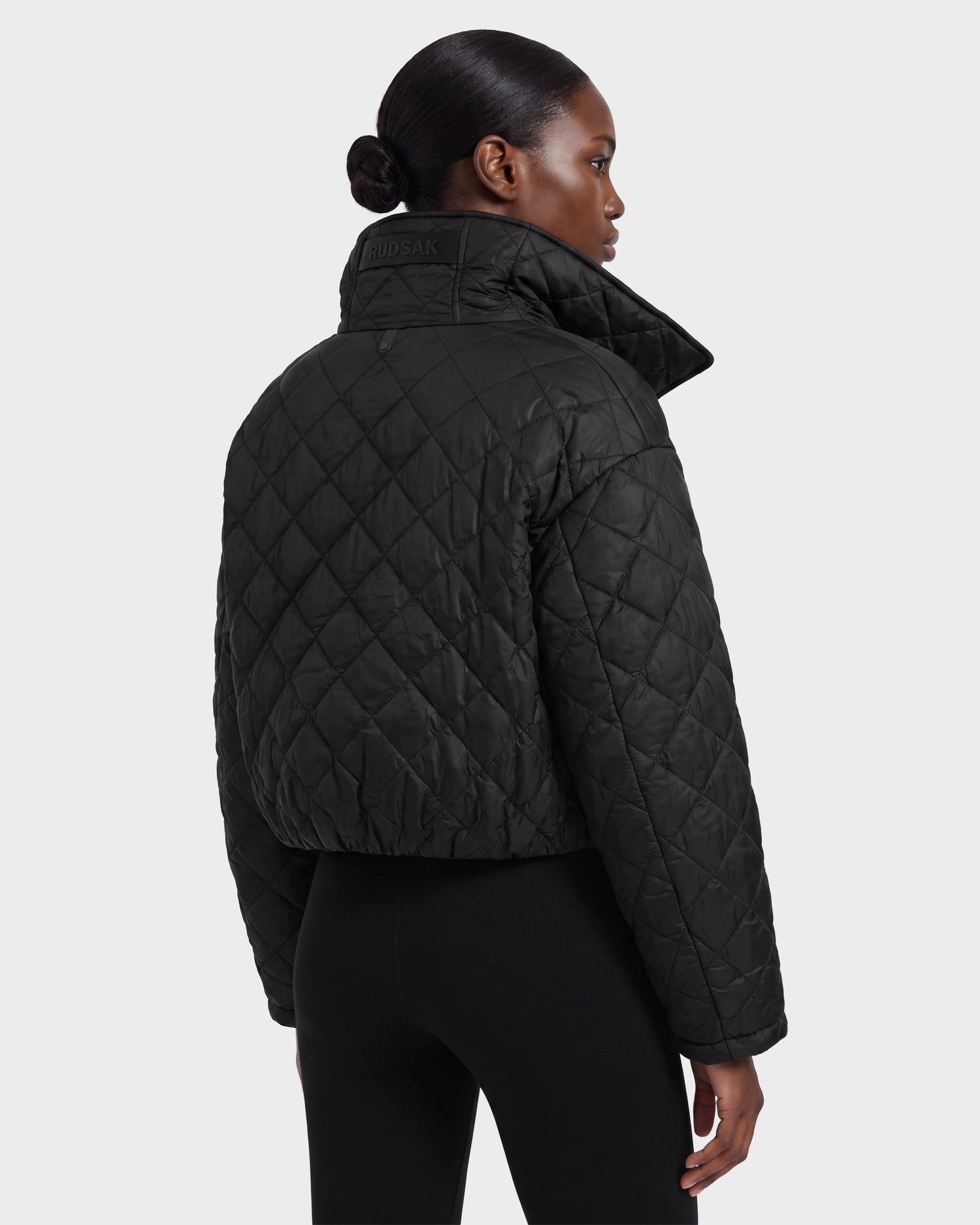 A person with neatly tied hair wears the Rudsak KALEE - 8124005 Black women's quilted light jacket—styled as a cropped black puffer—paired with black pants, standing against a plain white background and facing away from the camera. This look showcases active versatility while keeping a sleek, modern silhouette.