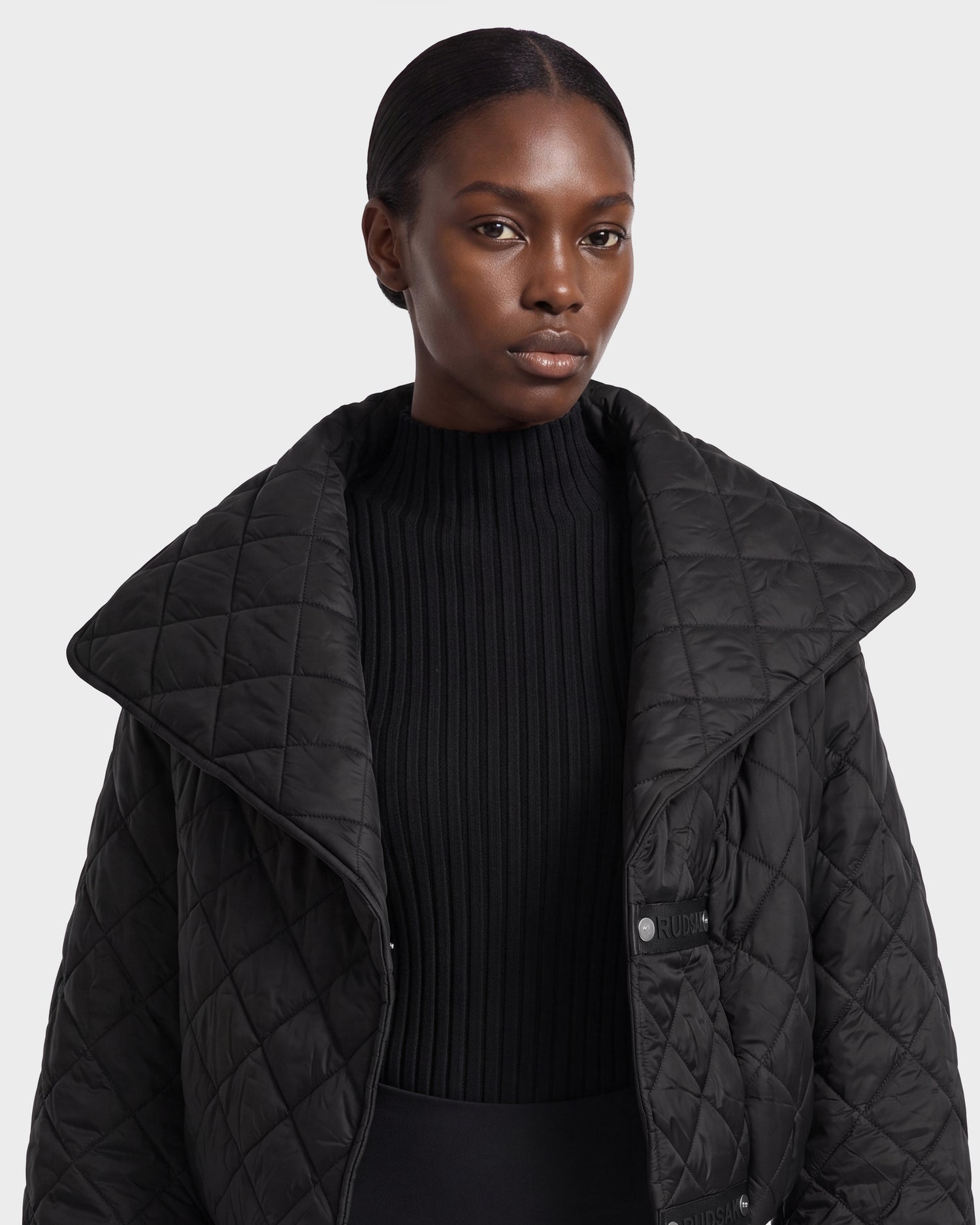 A woman with a serious expression wears the Rudsak KALEE - 8124005 Black quilted light jacket over a ribbed black turtleneck, showcasing its active versatility against a plain light background.