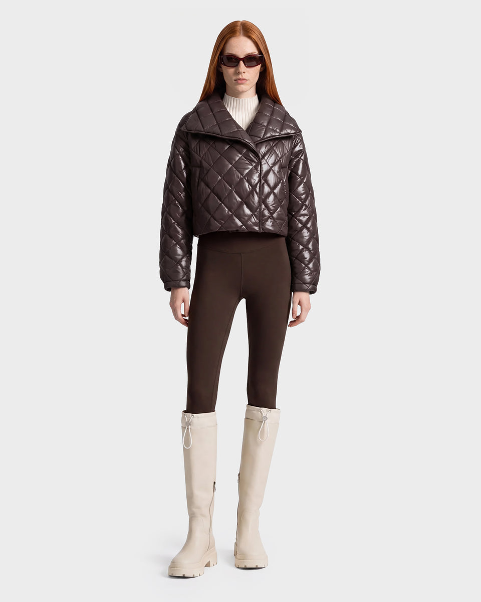A person wears sunglasses, a Rudsak KALEE - 8124005 Espresso brown quilted cropped jacket, dark leggings, a cream turtleneck, and tall off-white boots, standing against a plain light background.