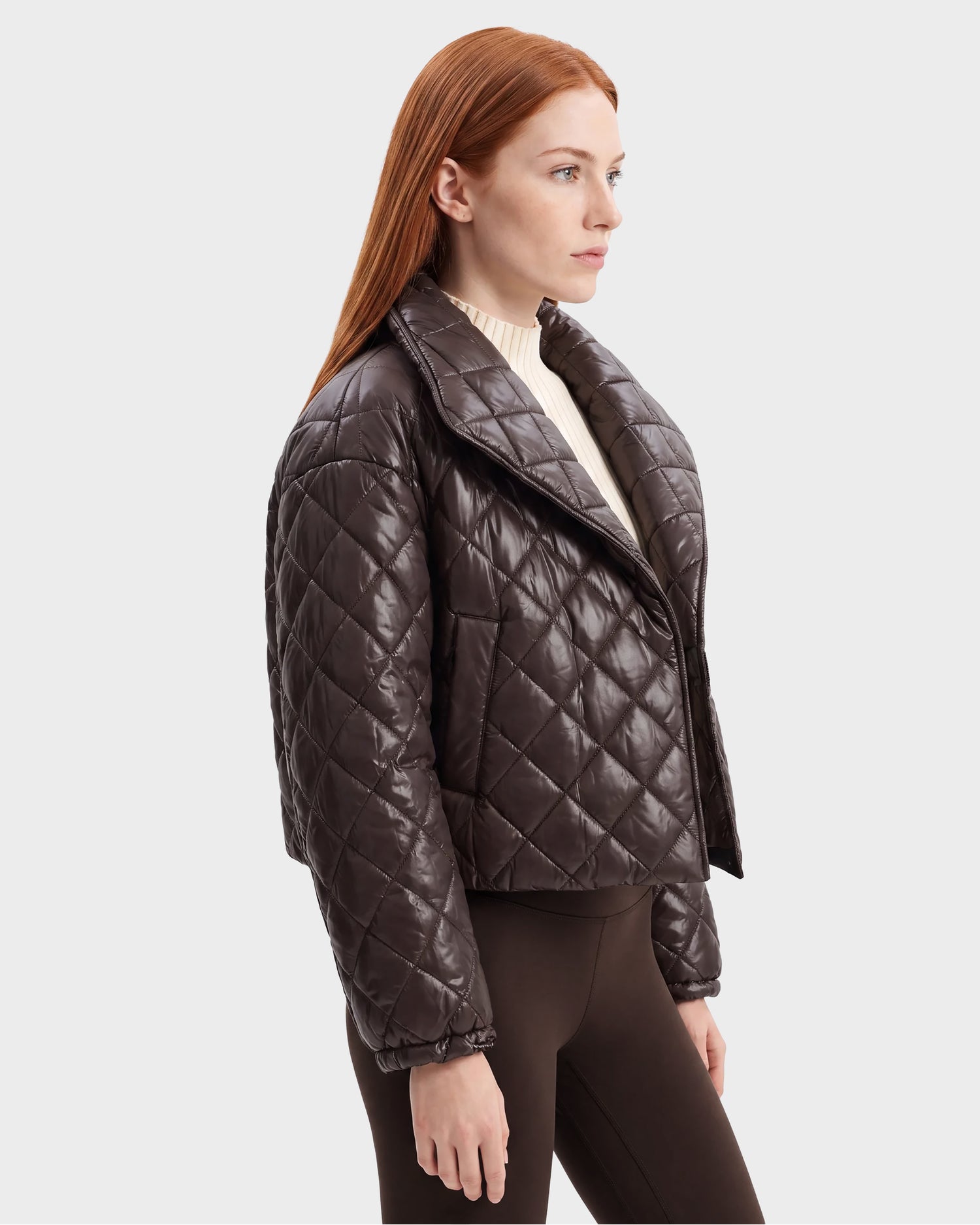 A woman with long red hair is wearing the Rudsak KALEE - 8124005 Espresso, a shiny dark brown women’s quilted light jacket, over a cream turtleneck and dark pants, standing in profile against a plain light background.