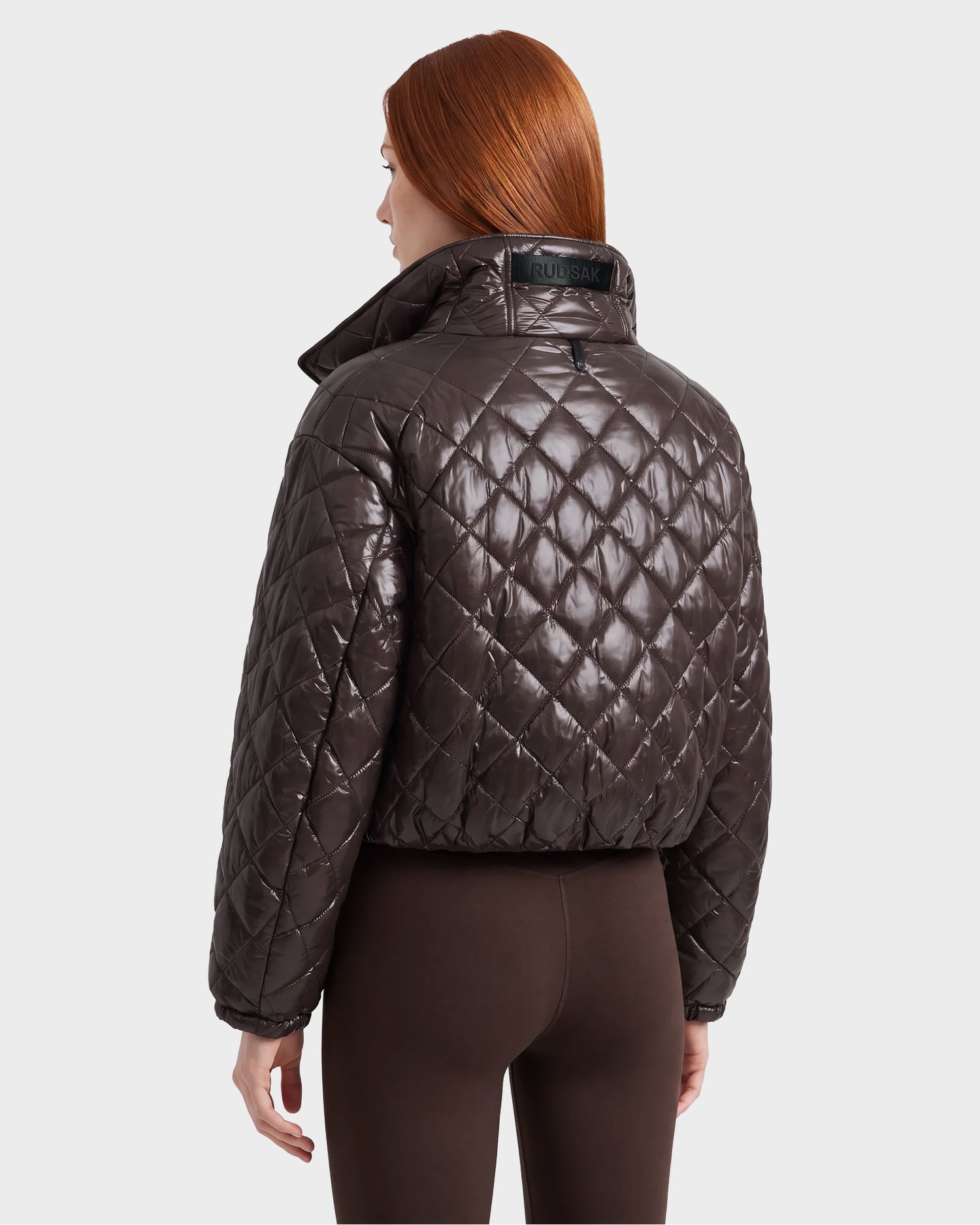 A person with long red hair is seen from behind wearing the Rudsak KALEE – 8124005 Espresso women’s quilted light jacket and dark pants, standing against a plain light background.