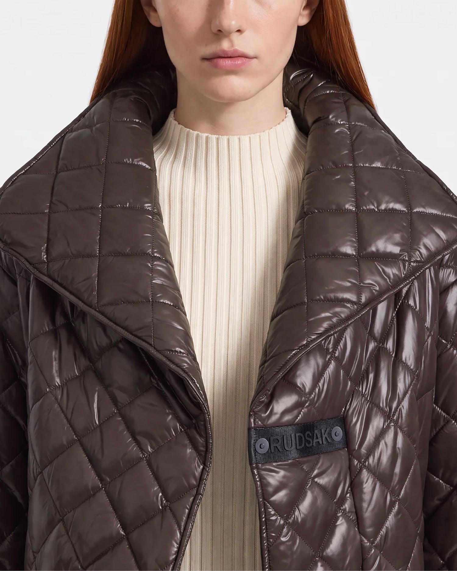 A woman with long reddish hair wears a shiny espresso-colored KALEE - 8124005 quilted light jacket by Rudsak, which features a large collar and a visible RUDSAK patch on the left side, over a cream ribbed top. The image captures her from the shoulders to mid-chest.