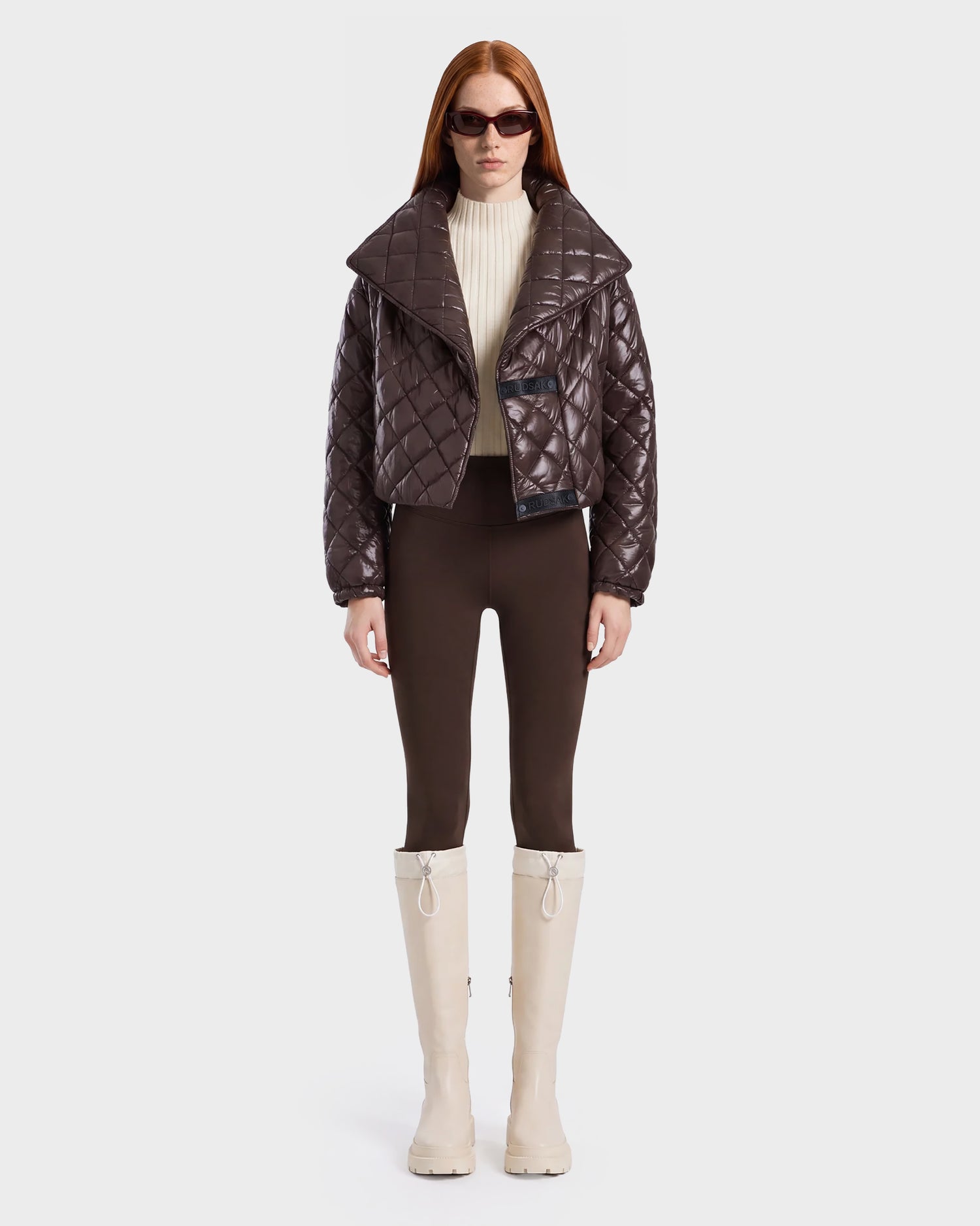 A woman with long red hair wears dark sunglasses, a shiny espresso Rudsak KALEE - 8124005 women's quilted light jacket, a cream turtleneck, brown pants, and tall white boots, standing against a plain white background.