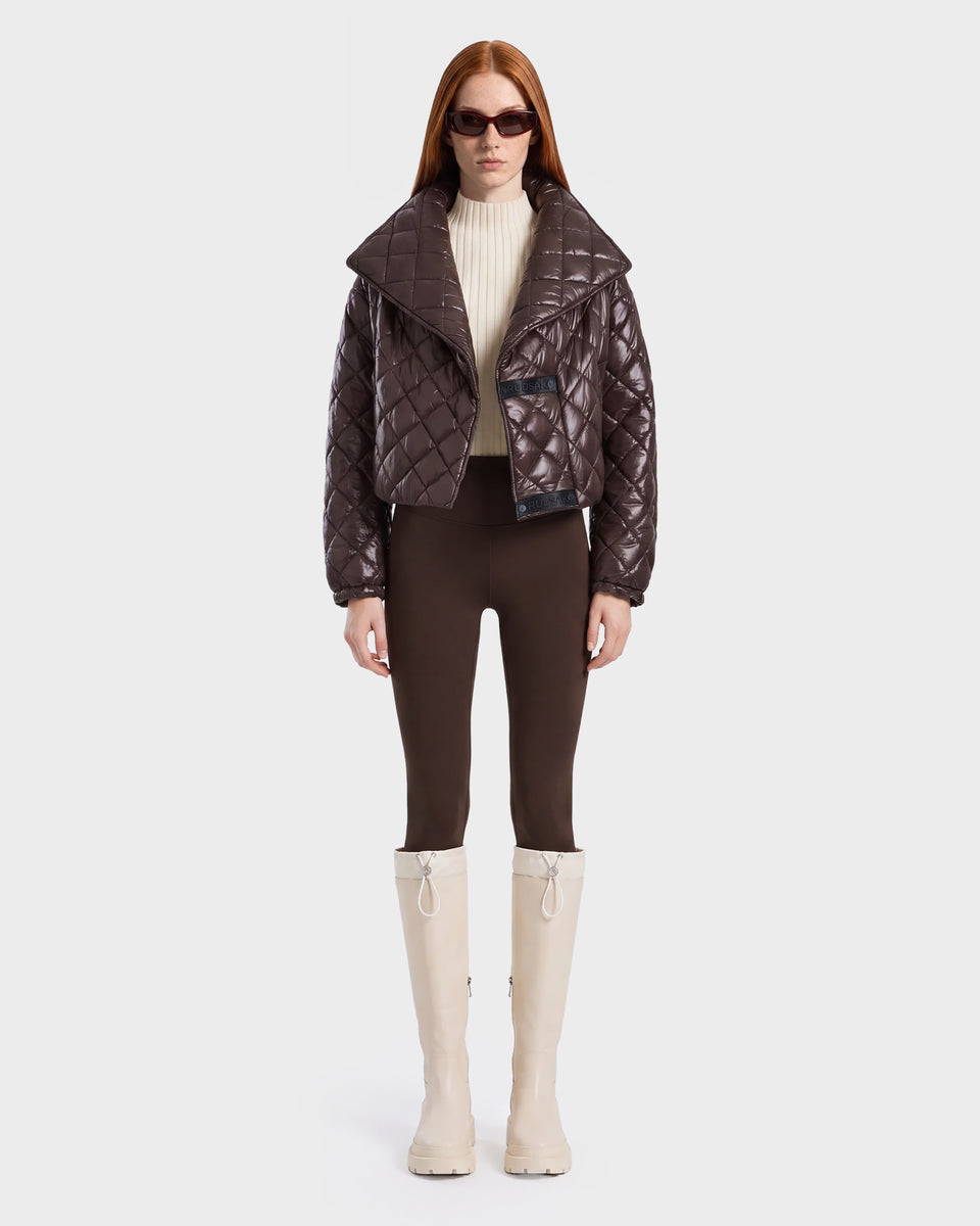 A woman with long red hair wears dark sunglasses, a shiny espresso Rudsak KALEE - 8124005 women's quilted light jacket, a cream turtleneck, brown pants, and tall white boots, standing against a plain white background.