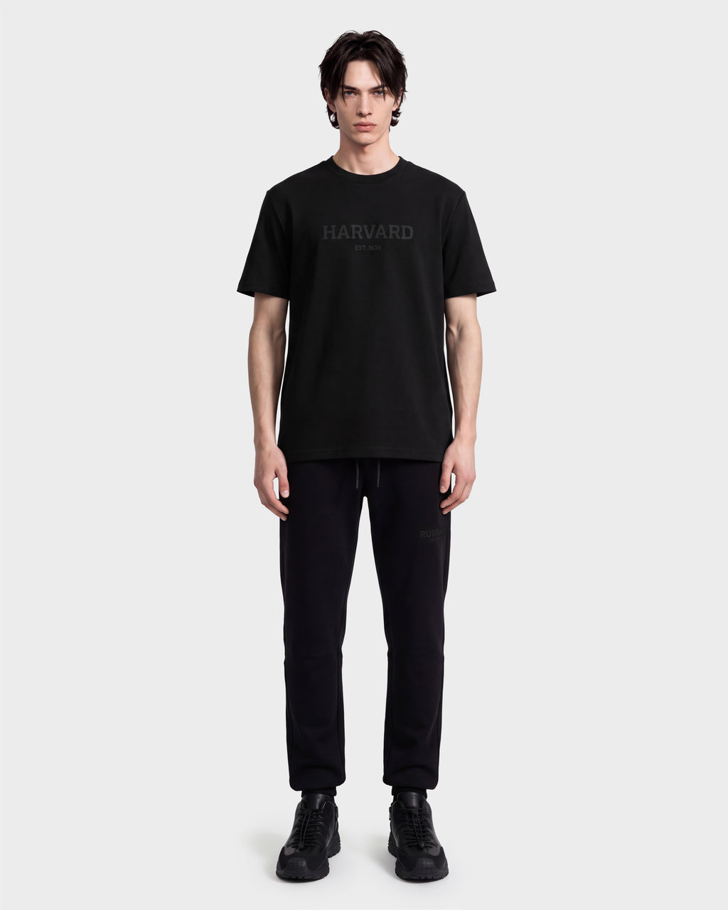 A person stands facing forward, wearing a black limited-edition Harvard apparel T-shirt, black pants, and KARTER R HV - H525507 Black shoes by Rudsak, against a plain white background.