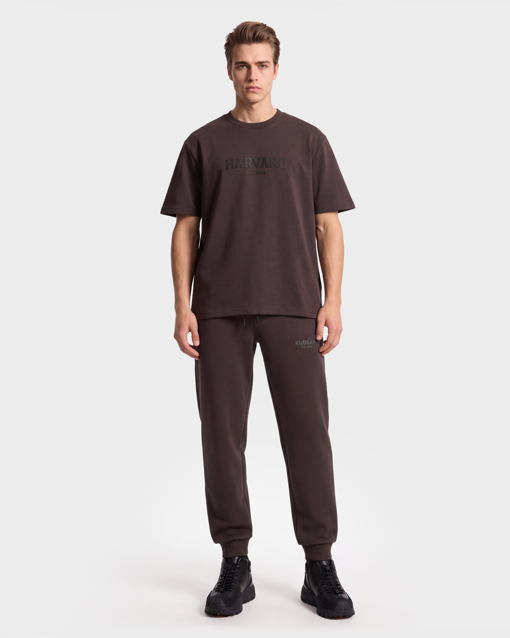A person stands facing forward, wearing the Rudsak KARTER R HV - H525507 Espresso limited edition T-shirt styled with matching jogger pants and black shoes, embracing a modern streetwear vibe against a plain white background.