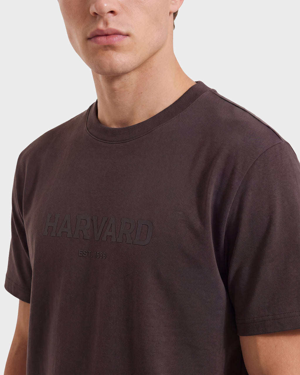 A person wearing the Rudsak KARTER R HV - H525507 Espresso limited edition Harvard T-shirt, featuring HARVARD EST. 1636 subtly printed on the front, is photographed from the shoulders up against a plain background, effortlessly channeling modern streetwear style.