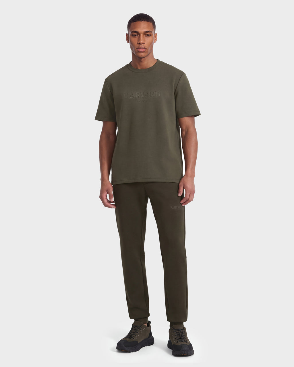 A man stands facing forward against a plain white background, wearing the Rudsak KARTER R HV - H525507 Olive streetwear t-shirt with matching jogger pants and black sneakers.