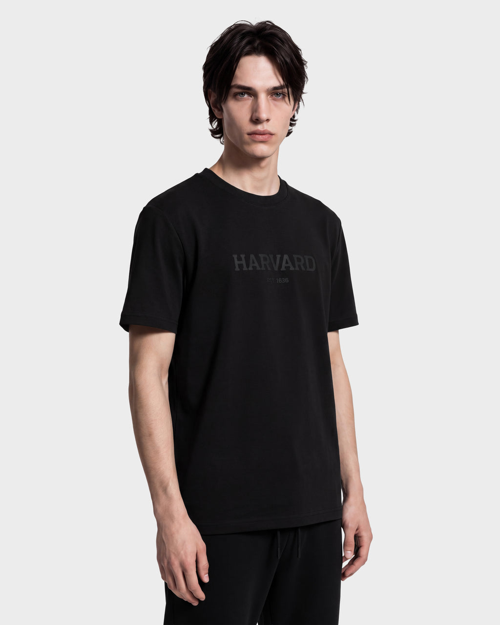 A young man with medium-length dark hair wears the KARTER R HV - H525507 Black limited-edition T-shirt by Rudsak, featuring HARVARD printed in dark letters across the chest. Showcasing a stylish blend of Rudsak’s Harvard apparel and streetwear, he stands against a plain white background, looking directly at the camera.