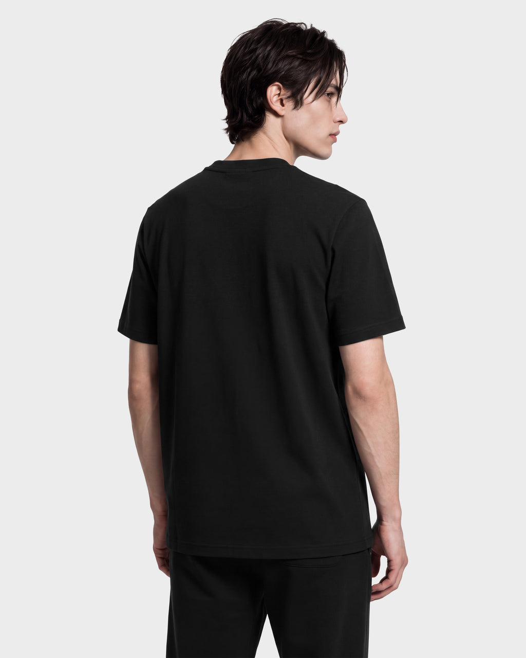 A person with short dark hair, wearing Rudsak’s KARTER R HV - H525507 Black limited-edition T-shirt and black pants, stands facing away and looks over their left shoulder against a plain light background.