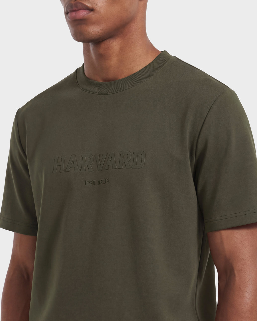 A person wearing the Rudsak KARTER R HV - H525507 Olive, a limited edition olive green Harvard T-shirt featuring “HARVARD EST. 1636” embossed on the front. The image highlights the upper body against a plain background, showcasing this distinctive piece from Rudsak’s streetwear collection.