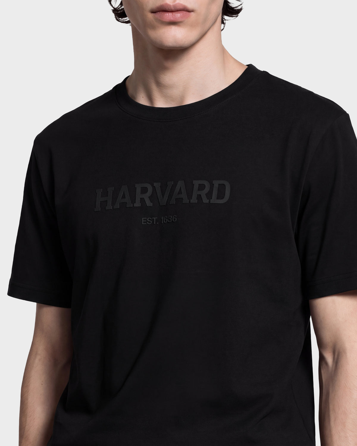 A person wearing the KARTER R HV - H525507 Black T-shirt by Rudsak, a limited-edition piece featuring HARVARD and EST. 1636 embossed on the front. This stylish upper-torso look—set against a plain background—highlights classic streetwear reimagined with an Ivy League edge, making it perfect for anyone looking to elevate their Harvard apparel collection.
