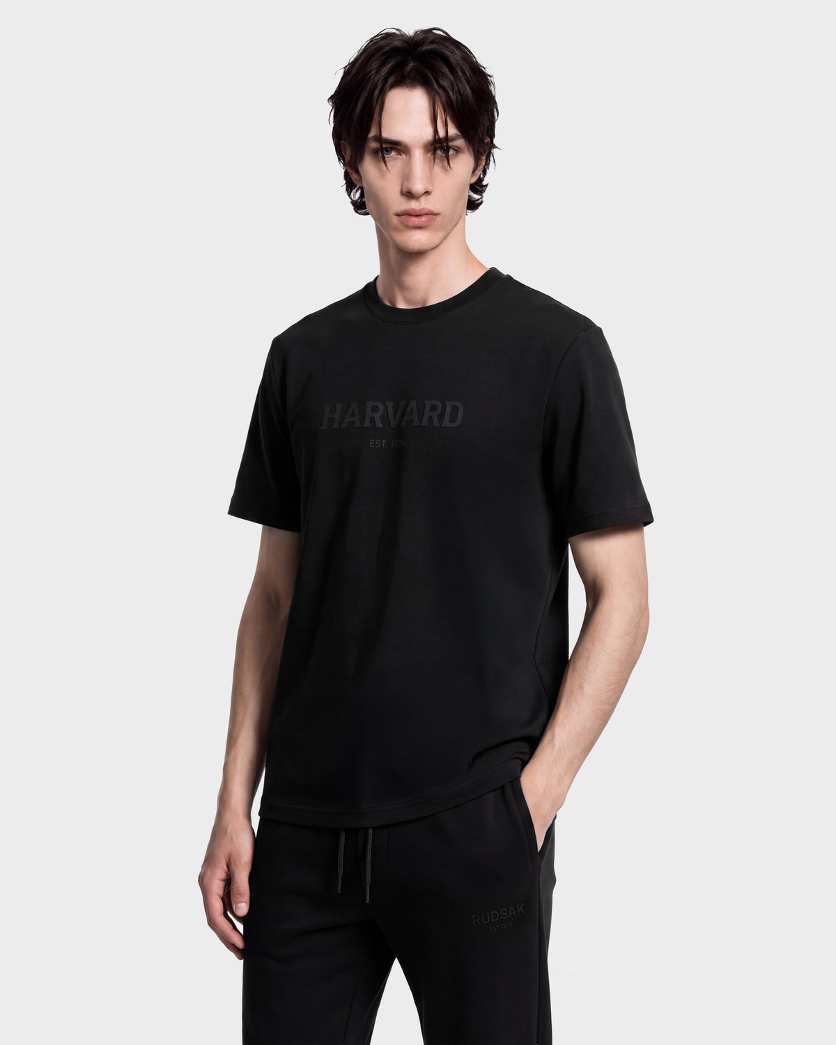 A young man with dark hair wears a limited-edition Harvard T-shirt and Rudsak KARTER R HV - H525507 black pants, posing with one hand in his pocket against a plain white background.