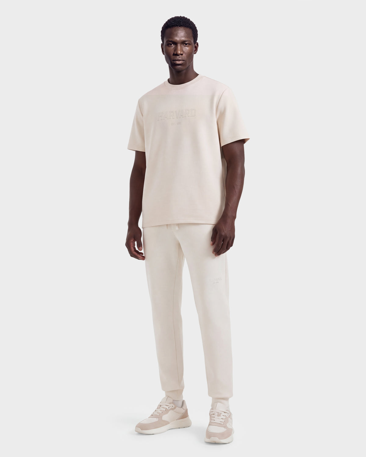 A man stands wearing the KARTER R HV - H525507 Vanilla limited edition short-sleeve shirt and joggers by Rudsak, paired with light beige sneakers, posing against a plain light background. His look channels laid-back streetwear vibes, effortlessly blending comfort and style in this signature set.