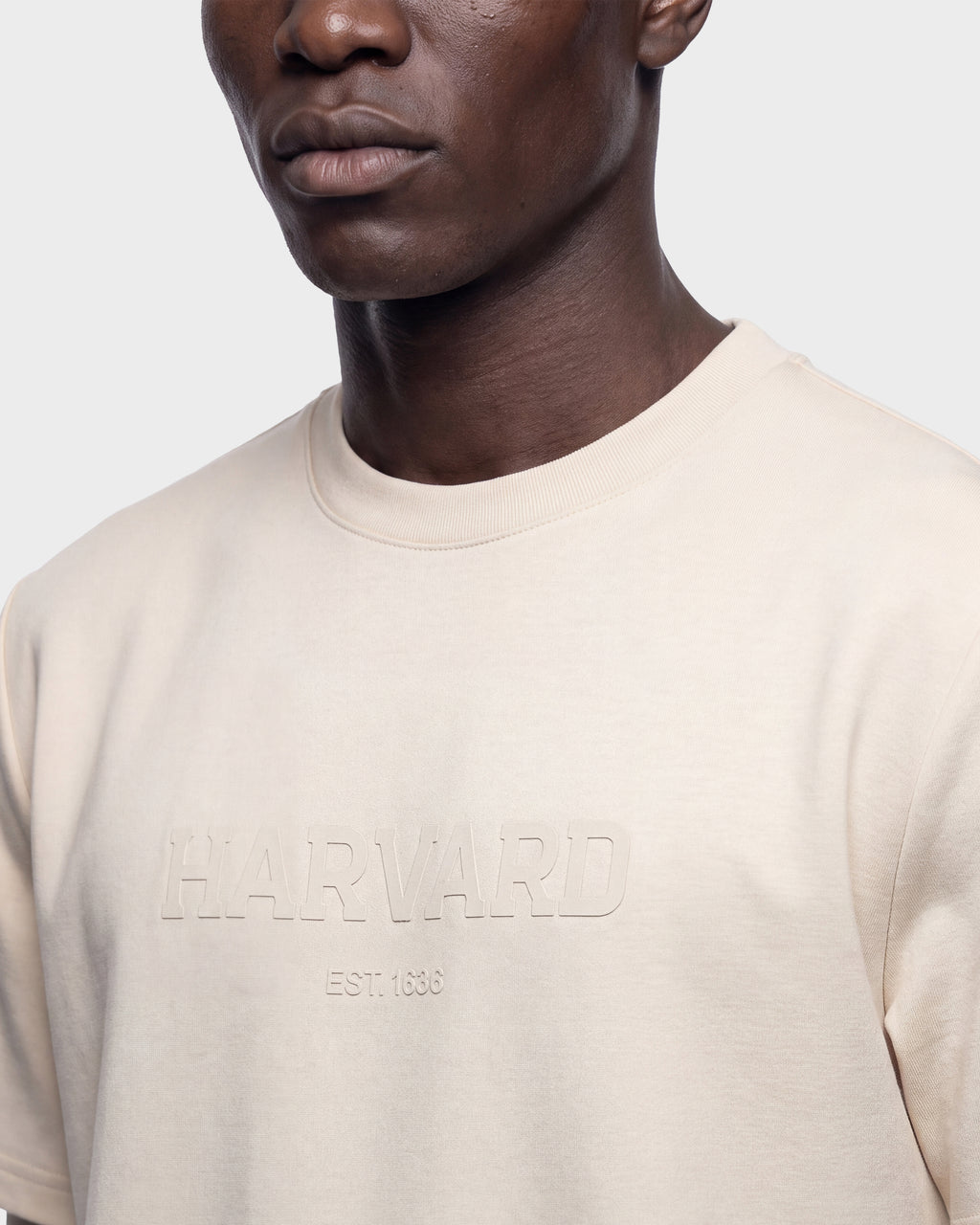 A person wearing the KARTER R HV - H525507 Vanilla T-shirt by Rudsak, featuring a light beige color and subtle embossed branding on the front. The image captures their upper torso and a partial view of their face against a plain background, creating a clean and modern streetwear-inspired aesthetic.