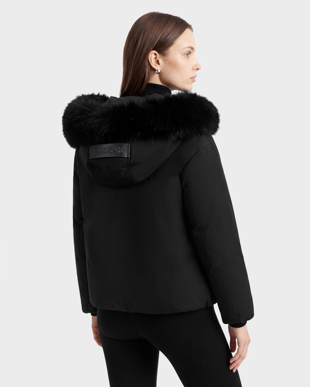 A woman wearing a black Rudsak KATHLEEN - 8126764 short recycled nylon puffer jacket with a fur-trimmed hood and a logo patch on the back.