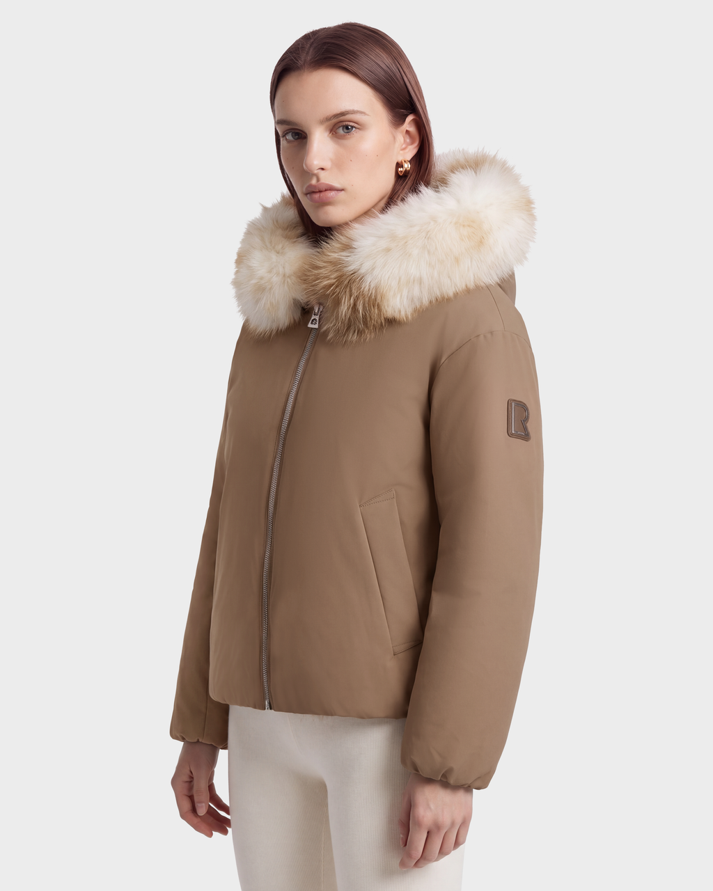 A person wearing a tan Rudsak KATHLEEN - 8126764 Sand hooded winter bomber jacket with a large faux-fur trimmed hood and a zippered front.
