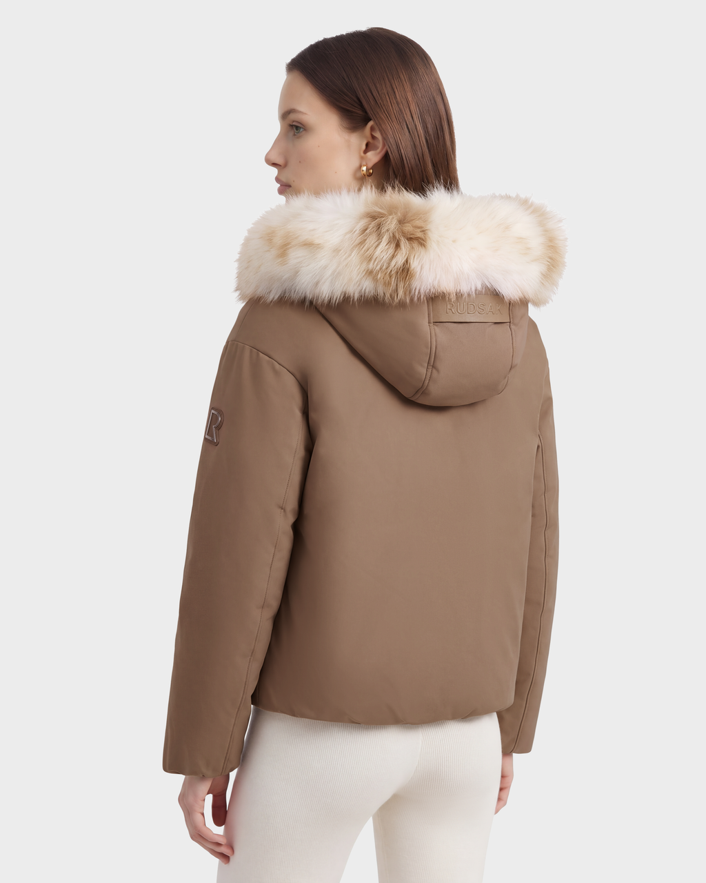 A person wears a tan Rudsak KATHLEEN - 8126764 Sand duck down-insulated puffer jacket featuring a hood with light-colored faux-fur detailing.