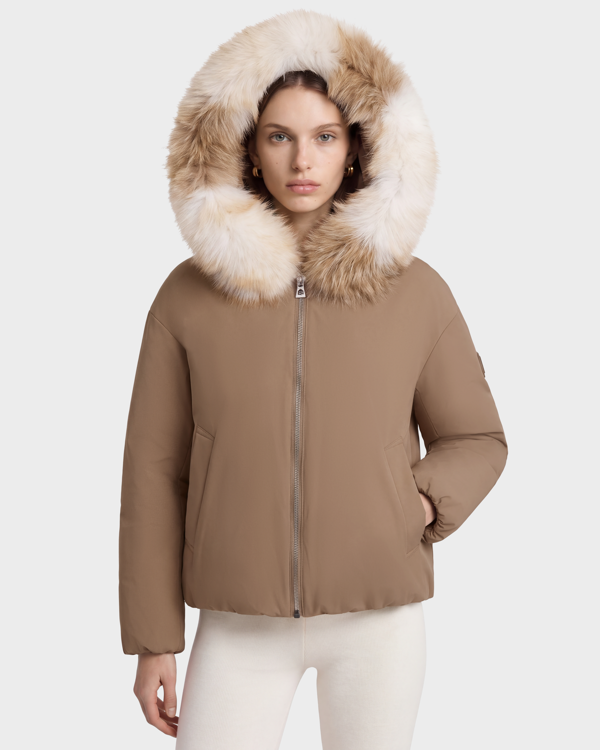 A woman wears a tan KATHLEEN - 8126764 Sand winter bomber jacket with a large faux-fur trimmed hood.