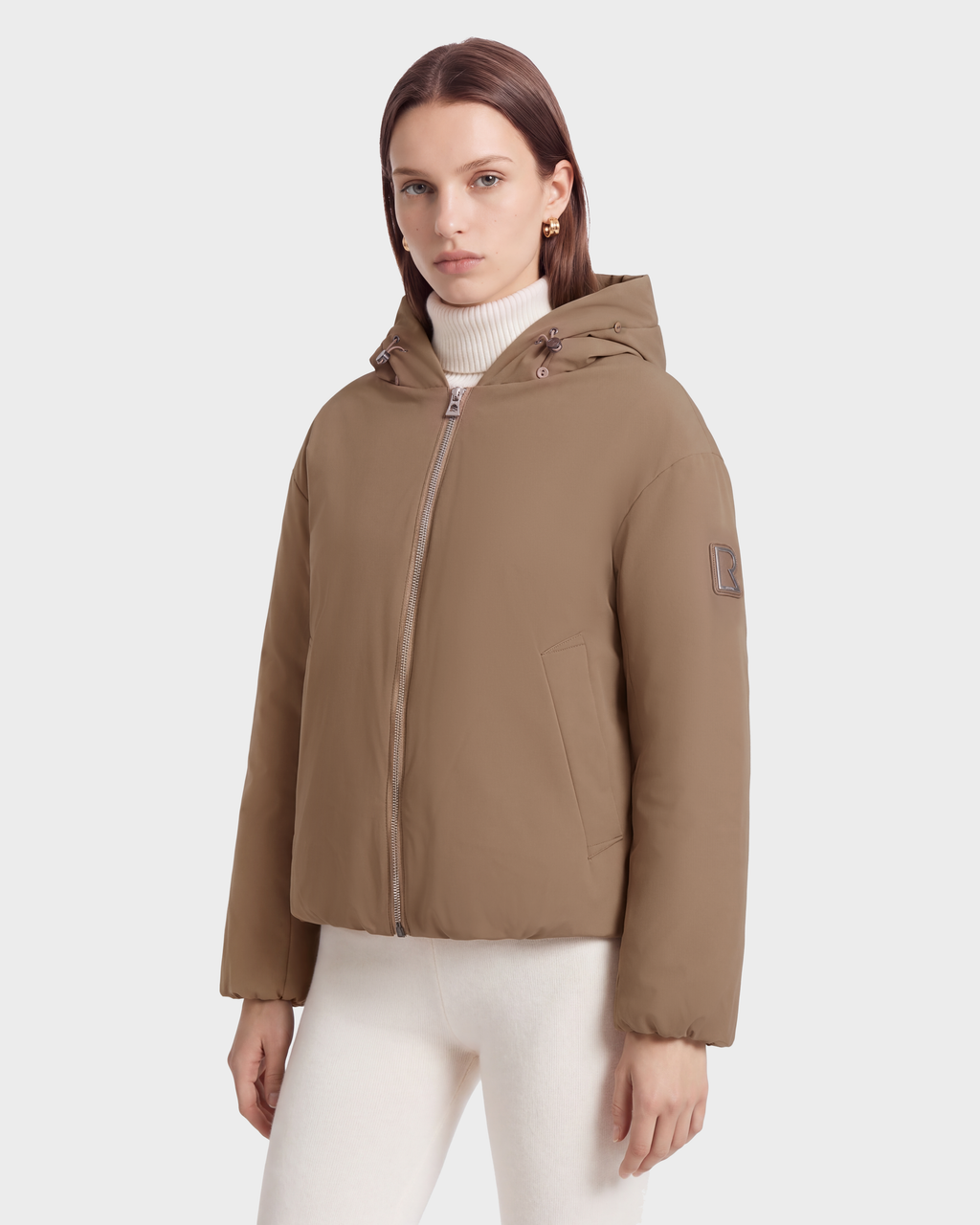 A person wearing a sand-colored, hooded, duck down-insulated KATHLEEN - 8126764 Sand puffer jacket over a cream-colored turtleneck.