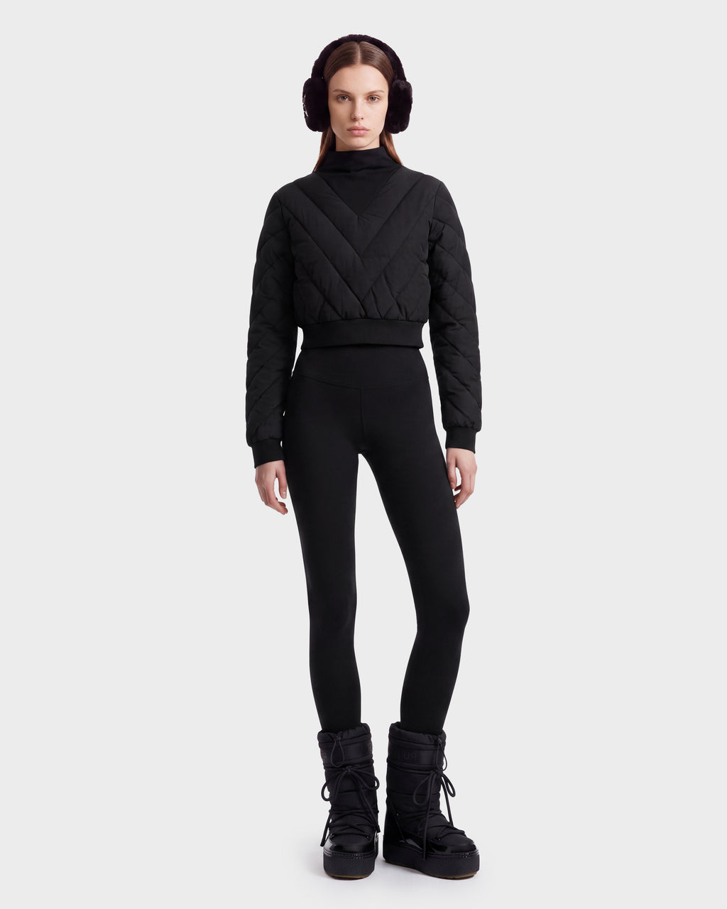 A person stands against a plain background wearing black earmuffs, sleek black leggings, and winter boots, styled with the Rudsak KATYA - 8526531 Black quilted sweater—perfect for an après-ski look.