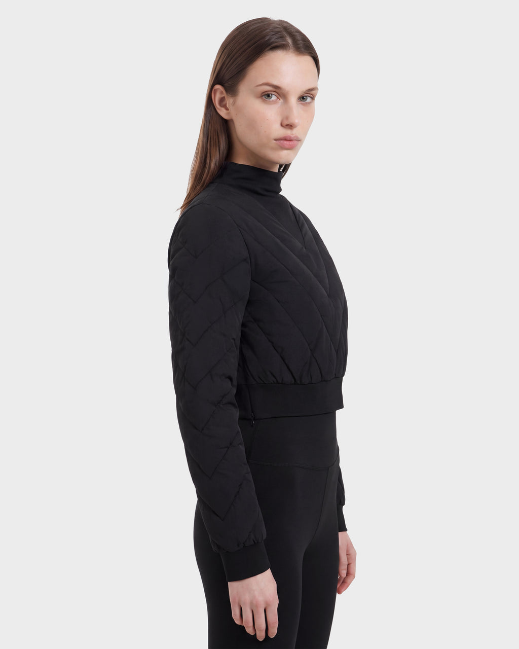 A person with straight brown hair stands against a plain light background, facing slightly to the side, wearing the KATYA - 8526531 Black quilted, long-sleeve cropped down jacket by Rudsak and black pants.