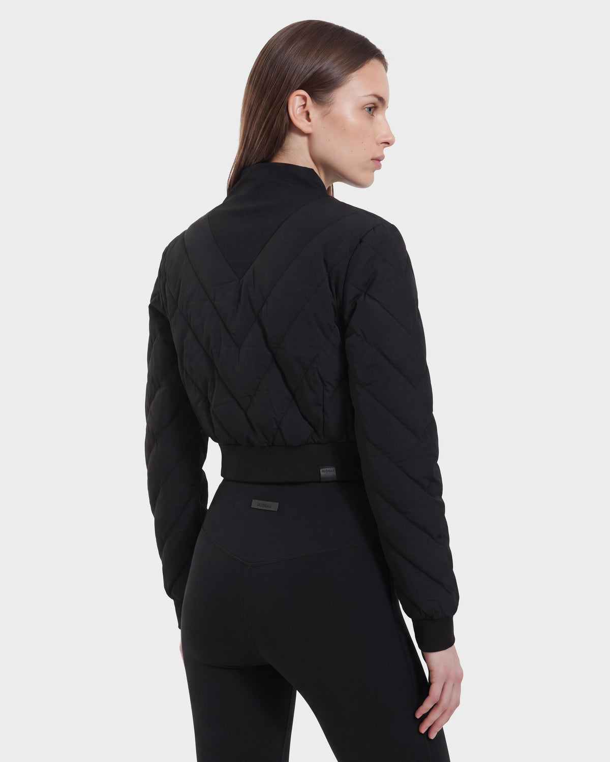 A woman with straight brown hair wears the Rudsak KATYA - 8526531 Black quilted down jacket and black pants, standing sideways and looking over her shoulder against a plain white background.