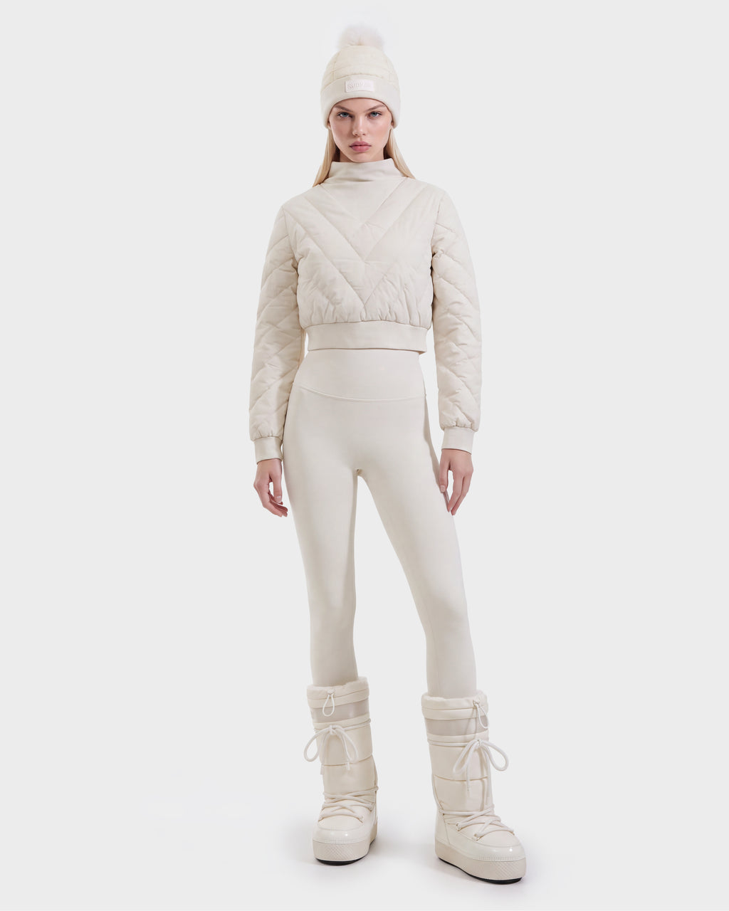 A woman is wearing the Rudsak KATYA - 8526531 Vanilla quilted down jacket, paired with matching leggings, a pom-pom beanie, and tall snow boots, standing against a plain white background.