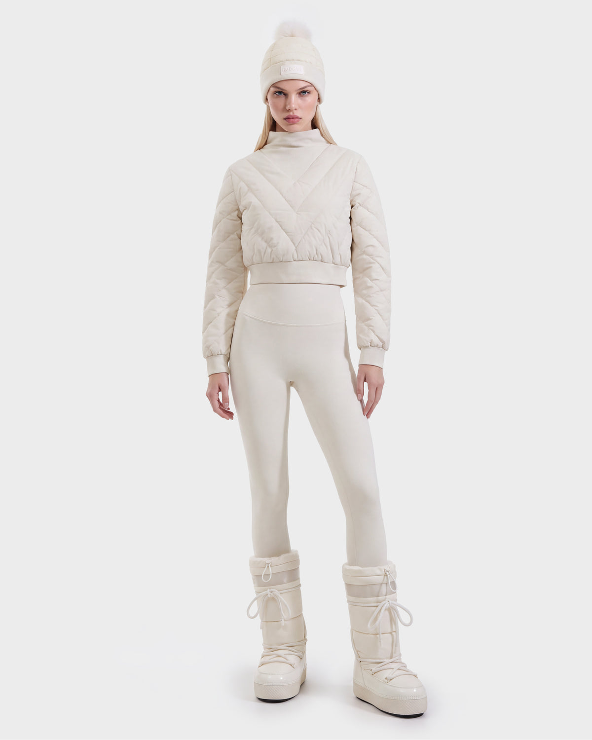 A woman is wearing the Rudsak KATYA - 8526531 Vanilla quilted down jacket, paired with matching leggings, a pom-pom beanie, and tall snow boots, standing against a plain white background.
