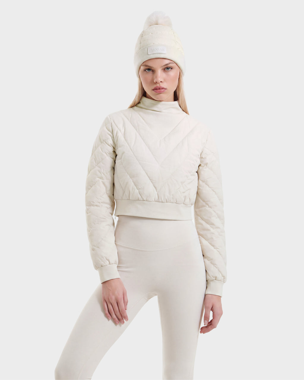 A woman models the Rudsak KATYA - 8526531 Vanilla cream-colored quilted down jacket, paired with matching high-waisted leggings and a knit beanie featuring a pom-pom, standing against a plain white background.