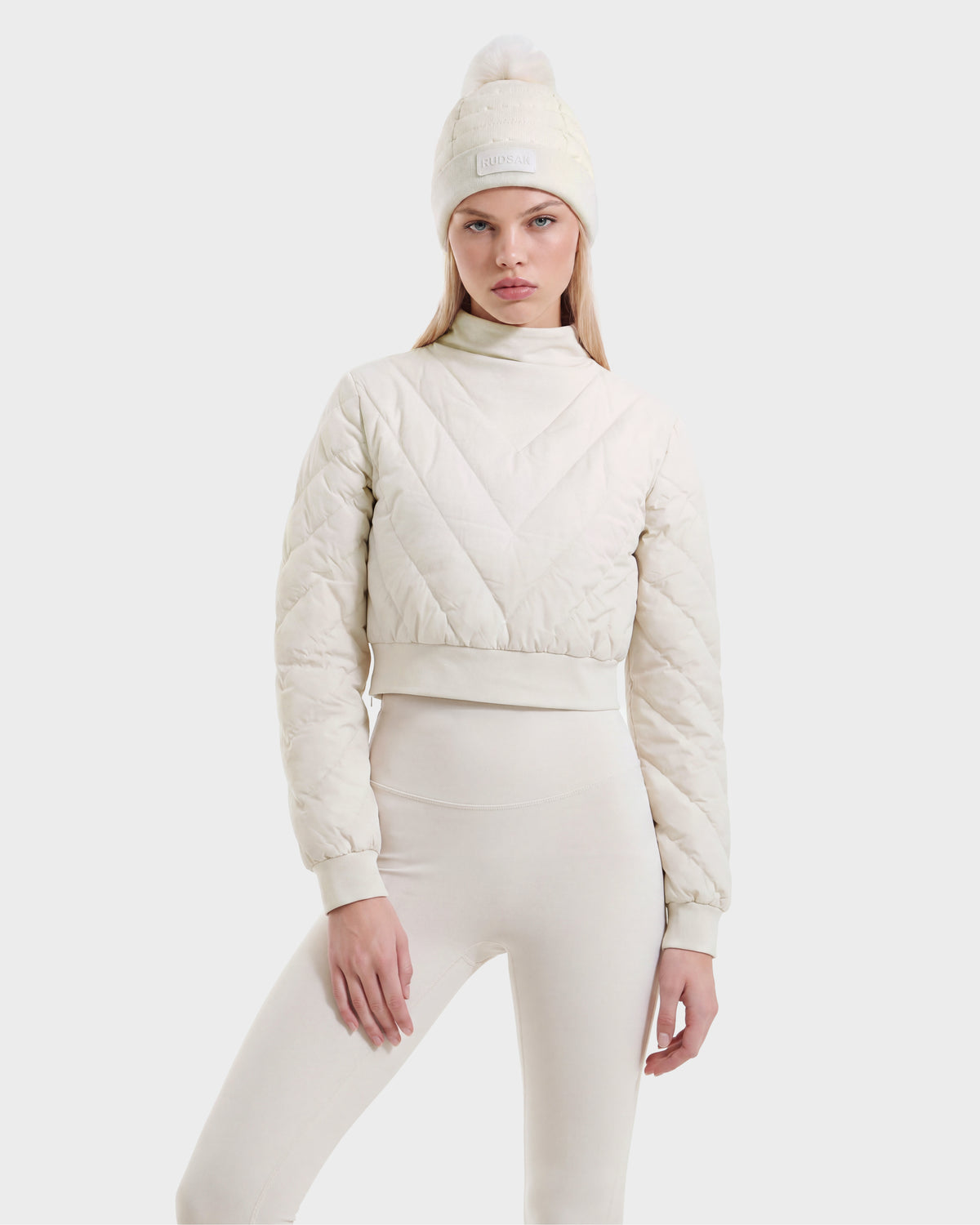 A woman models the Rudsak KATYA - 8526531 Vanilla cream-colored quilted down jacket, paired with matching high-waisted leggings and a knit beanie featuring a pom-pom, standing against a plain white background.