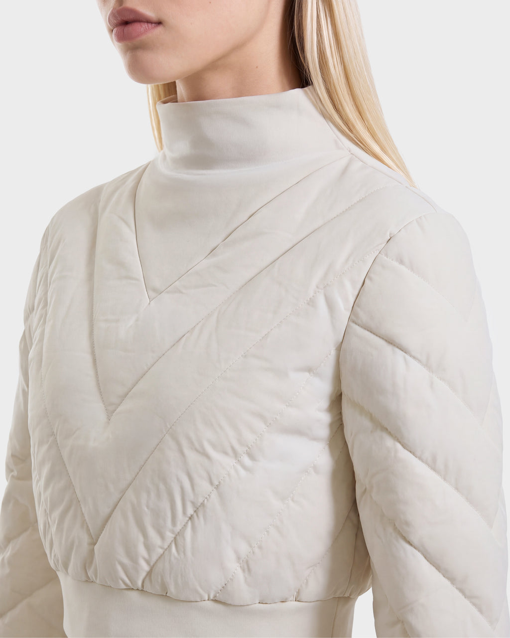 A woman with long blonde hair wears the Rudsak KATYA - 8526531 Vanilla, a cream-colored quilted top with down insulation, featuring a high collar and chevron pattern inspired by après-ski style. Only her upper body and face in partial profile are visible against a plain background.