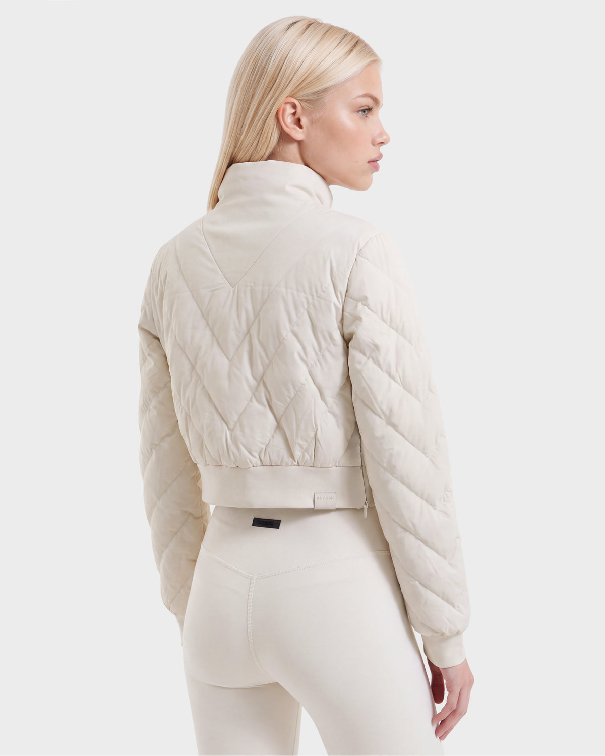 A woman with long blonde hair wears the Rudsak KATYA - 8526531 Vanilla quilted down jacket and matching leggings, standing with her back turned and looking to the side against a plain white background.