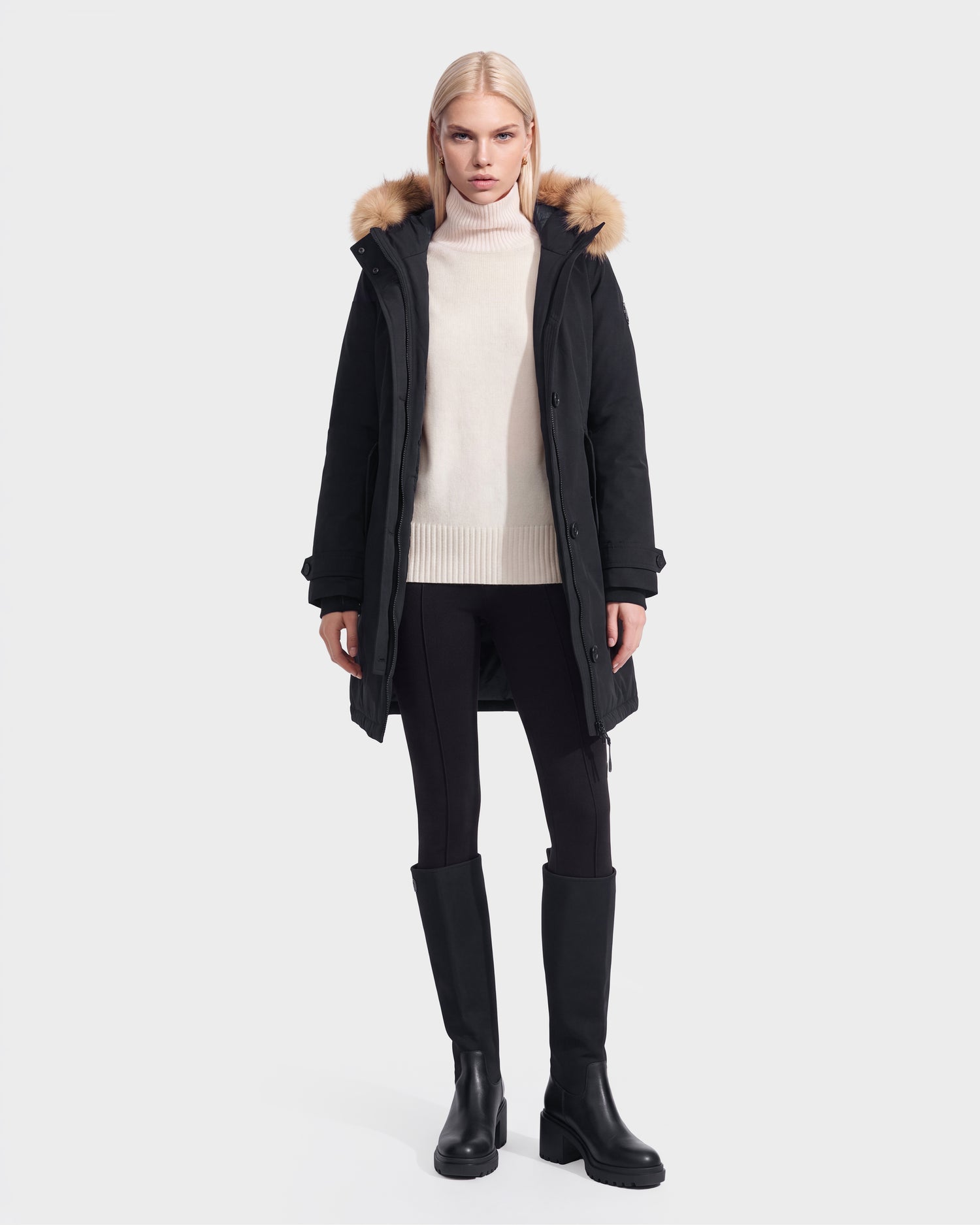 A person stands facing forward against a plain light background, wearing the Rudsak KEYSHIA - 8126517 Black parka with a fur-lined hood, a cream turtleneck sweater, black leggings, and black knee-high boots.
