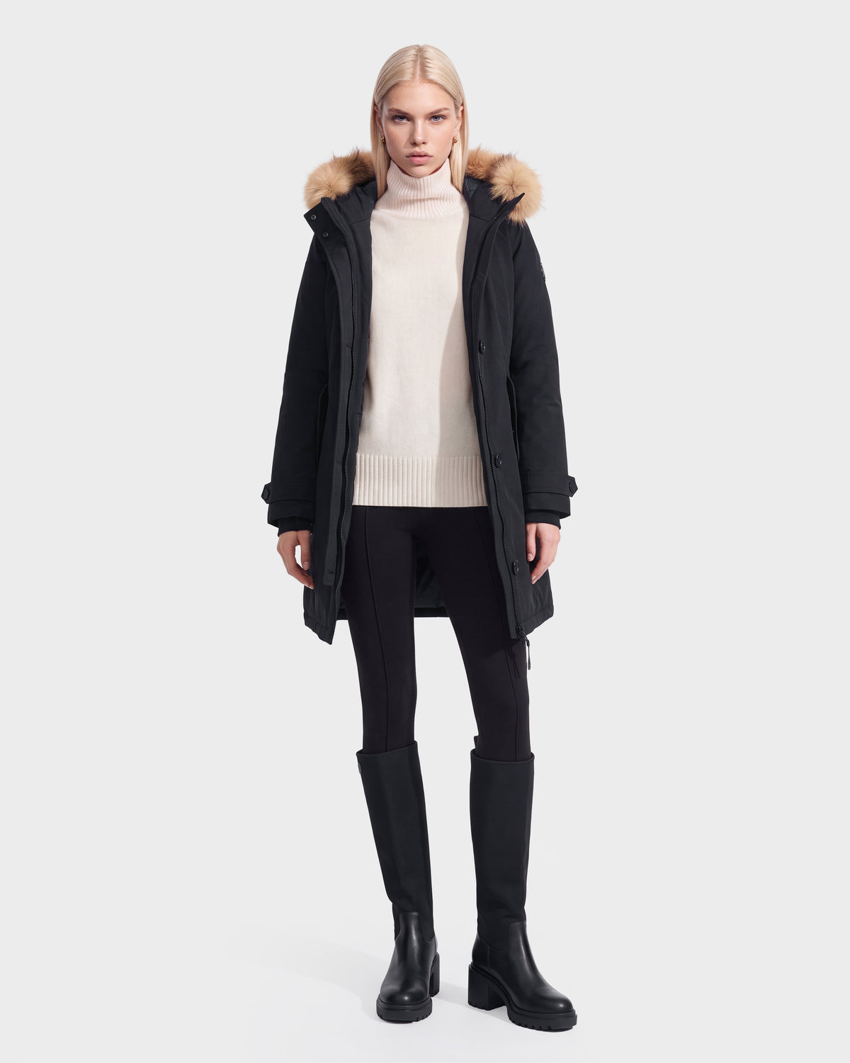 A woman models the Rudsak KEYSHIA - 8126517 Black down parka with a fur-trimmed hood, cream sweater, black leggings, and knee-high boots against a white background.